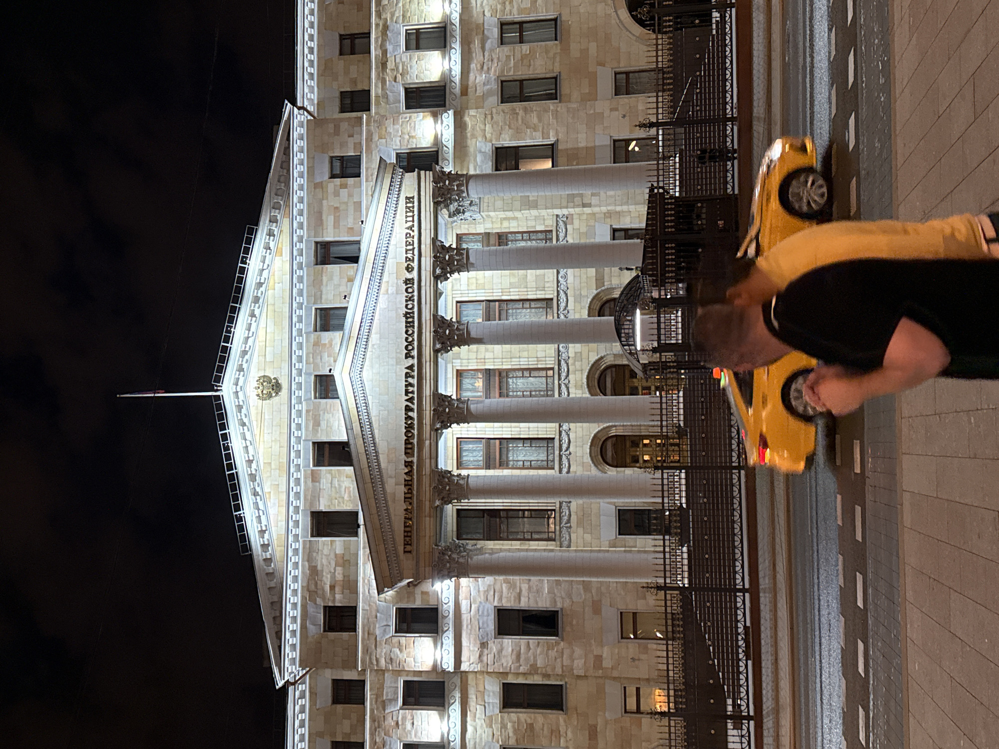 The image depicts the General Prosecutor's Office of the Russian Federation building at night in Moscow, Russia. The facade of the building is illuminated, featuring classical architecture with columns and ornate detailing. A Russian flag flies from a flagpole atop the building. In the foreground, a yellow taxi is driving past, and two individuals are walking on the sidewalk. The street is wet, reflecting the building's lights, and the sky is dark. The scene conveys a sense of urban nighttime activity with a prominent government building as the backdrop.
