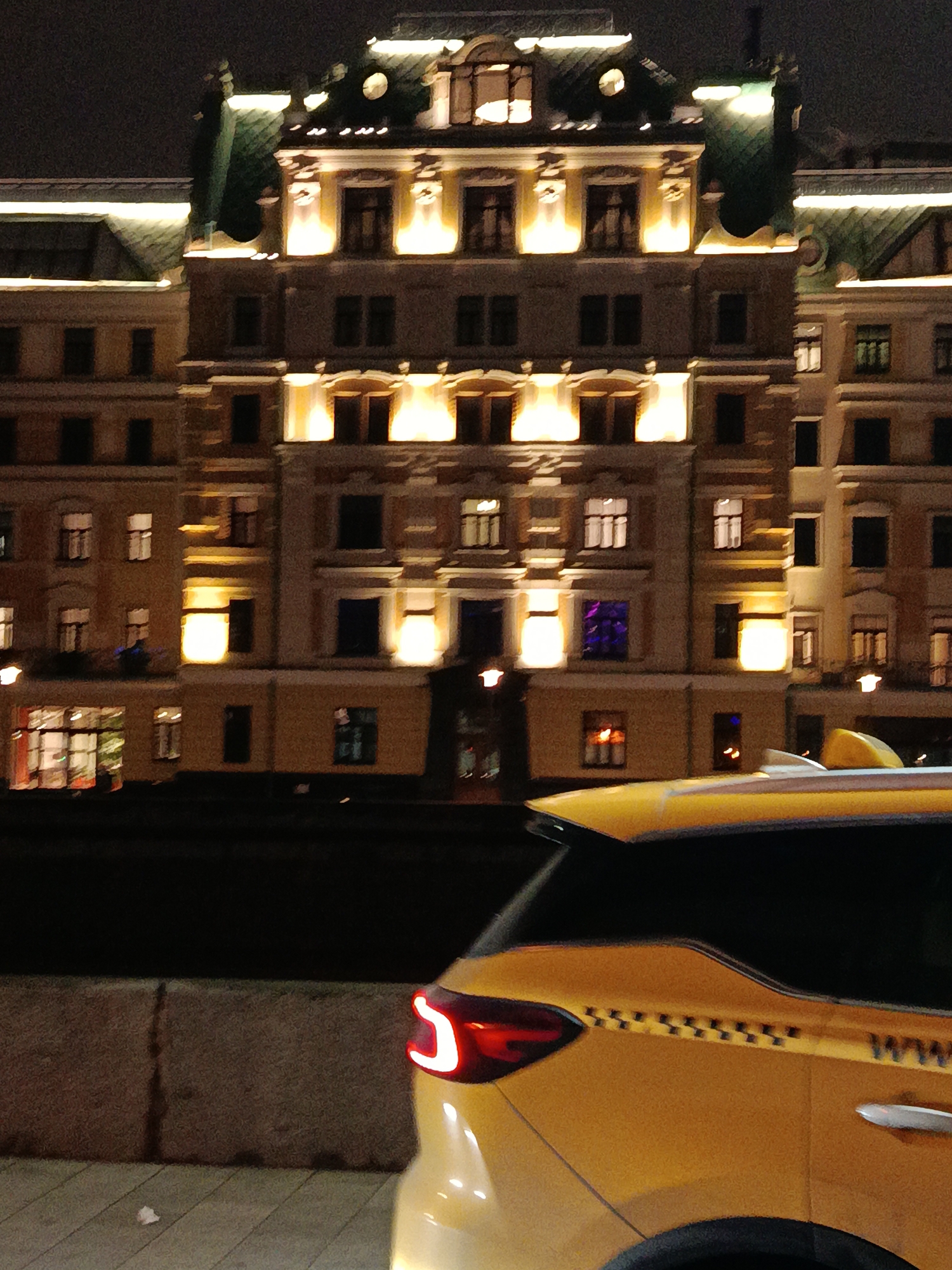 The image depicts a nighttime urban scene, focusing on a building illuminated by exterior lights and a yellow taxi in the foreground. The building is multi-storied with a facade featuring symmetrical windows and architectural details highlighted by the lighting. The taxi, partially in the frame, has a visible taillight and checkered pattern design, indicating its use as a taxi service. The environment appears to be a street or roadside, with elements such as a barrier wall suggesting an urban setting. The scene is likely in a city with a blend of modern and traditional architecture.