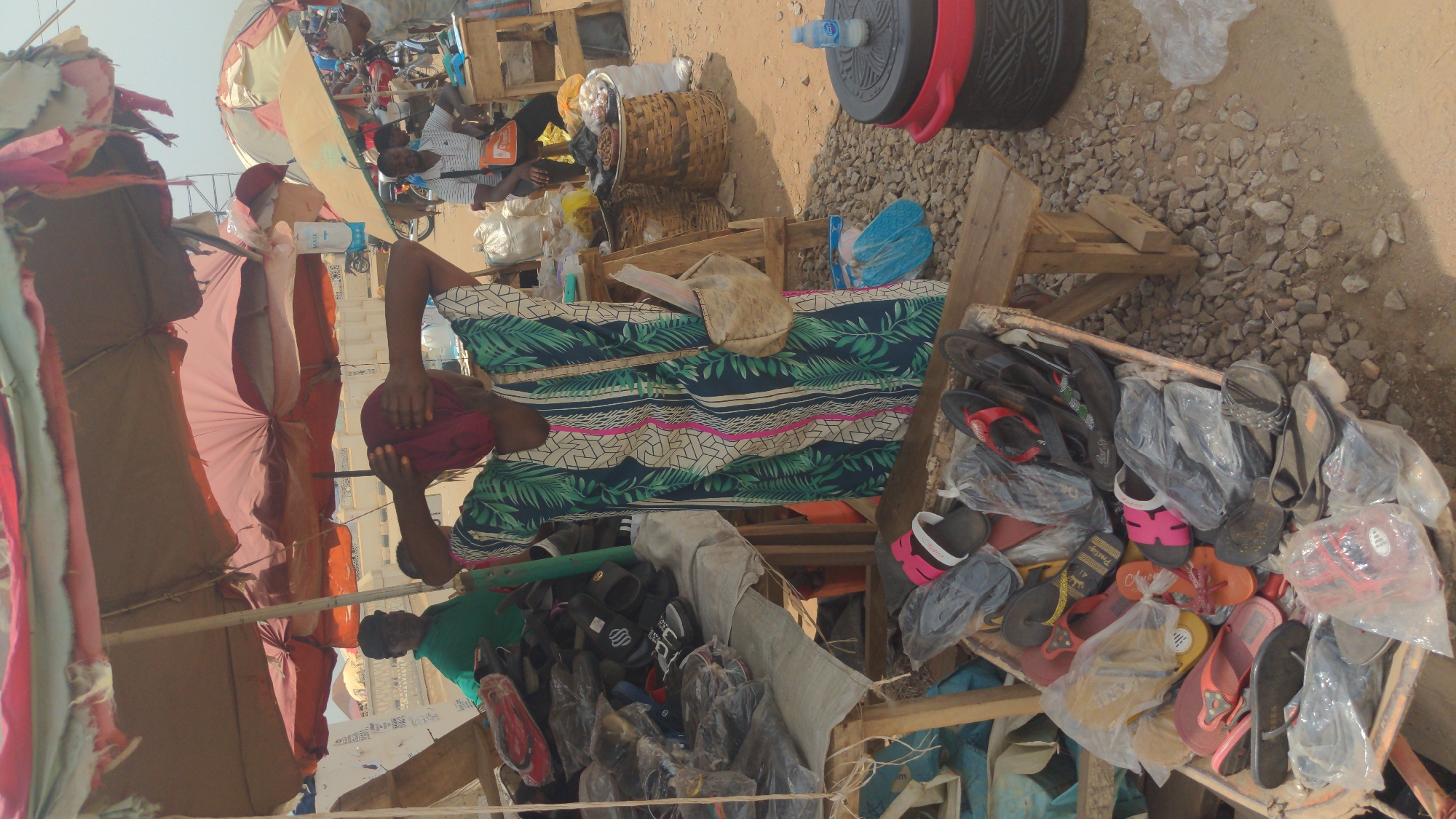 The image shows a street scene in Ado, Nigeria. A woman wearing a patterned dress and red headscarf sits behind a table covered with the same patterned fabric. In front of the table are wooden crates filled with flip-flops and sandals, some still in plastic bags. Behind her are other vendors and customers under awnings. A tire and a water jug are also visible near the table. The ground is rocky and sandy.