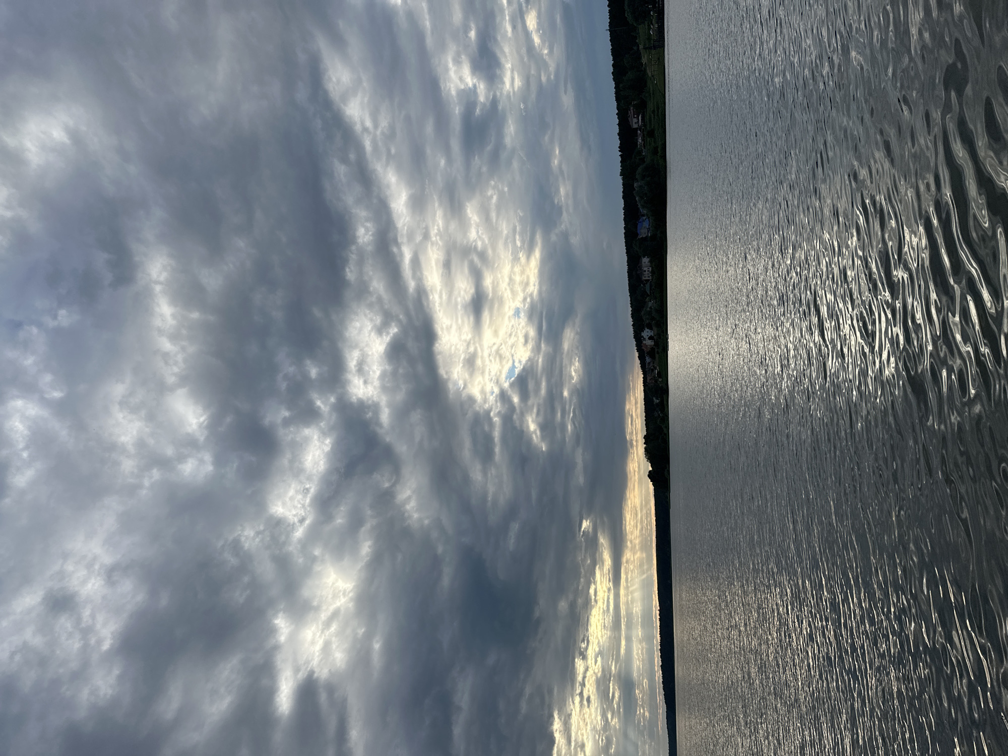 The image depicts a lake with a shoreline lined with trees and houses. The sky is filled with dramatic clouds, with the sun breaking through in some areas, creating rays of light. The water's surface is rippled, reflecting the sky and light. The scene evokes a sense of a serene evening or early morning in a natural setting, possibly a lakeside community. The time of day appears to be either sunrise or sunset, judging by the quality of light and the atmospheric conditions. The location context provided is Votkinsk, Russia. There is no visible text in the image.