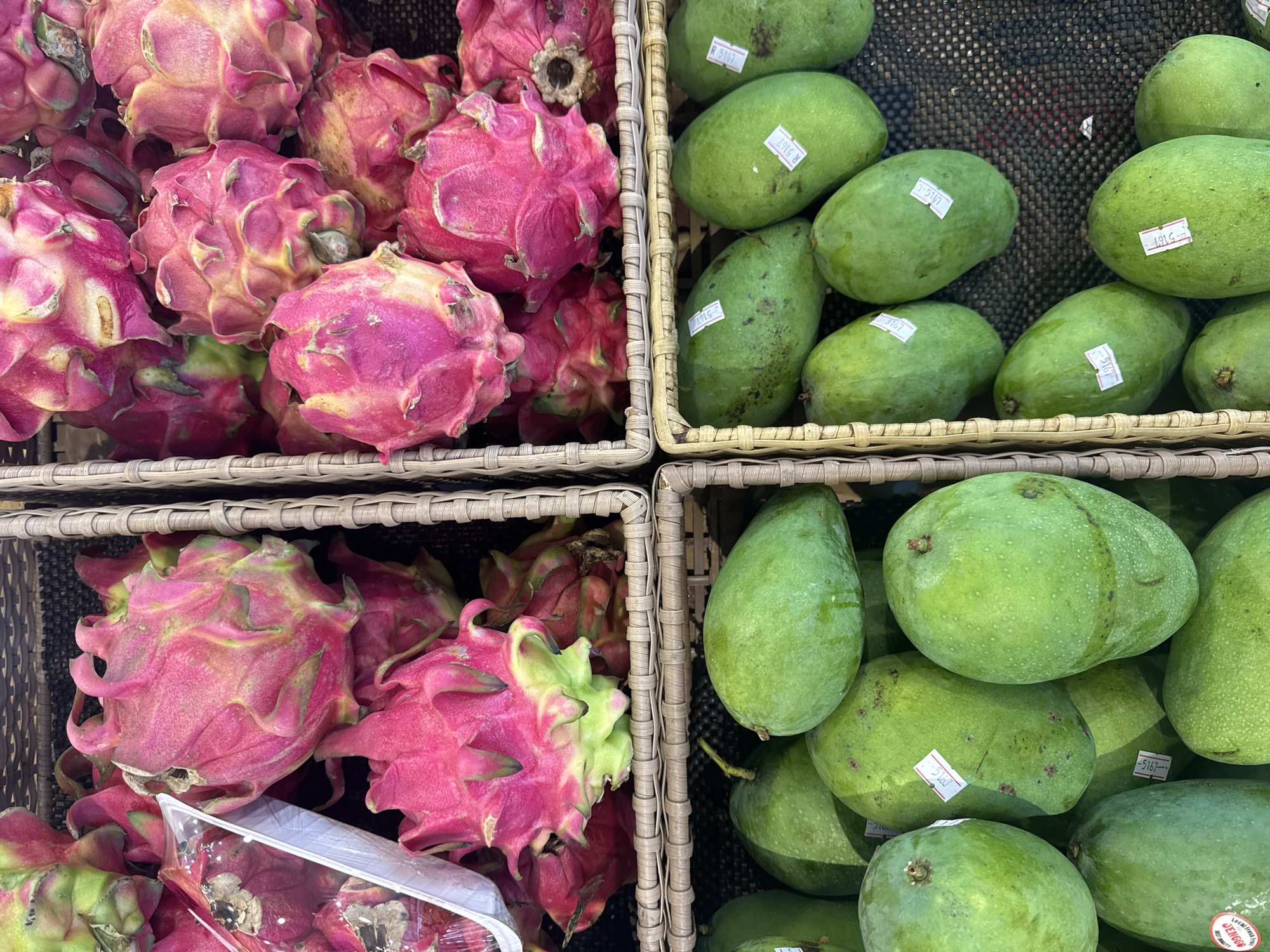 A high-angle shot depicts four woven baskets containing fruit. The left two baskets hold dragon fruit with pink skin and green protrusions. One of the baskets contains a wrapped package of dragon fruit. The right two baskets hold green mangoes with price tags. The location is implied by the presence of fruit sold at a market or shop in Kuta Selatan, Indonesia. No people are visible.