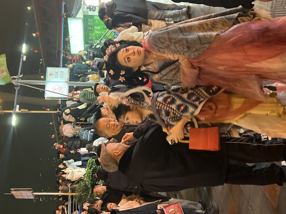 The image depicts a crowded nighttime urban scene in Yuzhong District, China, illuminated by artificial lights. In the foreground, two young women are prominently featured, dressed in elaborate traditional Chinese-style costumes.
The woman on the left wears a cream-colored gown with gold accents, a patterned blue, brown, and white shawl, and an intricate headdress with white and gold elements. She holds a white smartphone in her left hand and a small red paper shopping bag. She is looking towards an older man standing to her left. This older man wears a black coat and a plaid flat cap, and his right hand is raised near his chin as he appears to be speaking or interacting with the woman in costume. Standing slightly behind him and to the right is another older man, smiling and looking in the direction of the interaction.
The woman on the right wears a layered pink or reddish gown and a patterned grey and pink shawl. Her hair is styled in an elaborate updo adorned with multiple small pink flower-like decorations. Her gaze is directed towards the right, off-camera.
The background is densely populated with a crowd of people, many of whom are holding smartphones, suggesting photo-taking or recording. Some individuals are ascending or descending a set of stairs or an elevated platform. Overhead, bright streetlights illuminate the area. Visible signs include a white square sign with a green upward arrow and Chinese characters reading "出口" (Exit), alongside a red circle with a diagonal line indicating a prohibition. A green flag with white Chinese characters is affixed to a pole. In the deeper background, an elevated structure, possibly a bridge or overpass, is visible, marked with small red lights. Another green rectangular sign with white Chinese characters is present further back on the right.