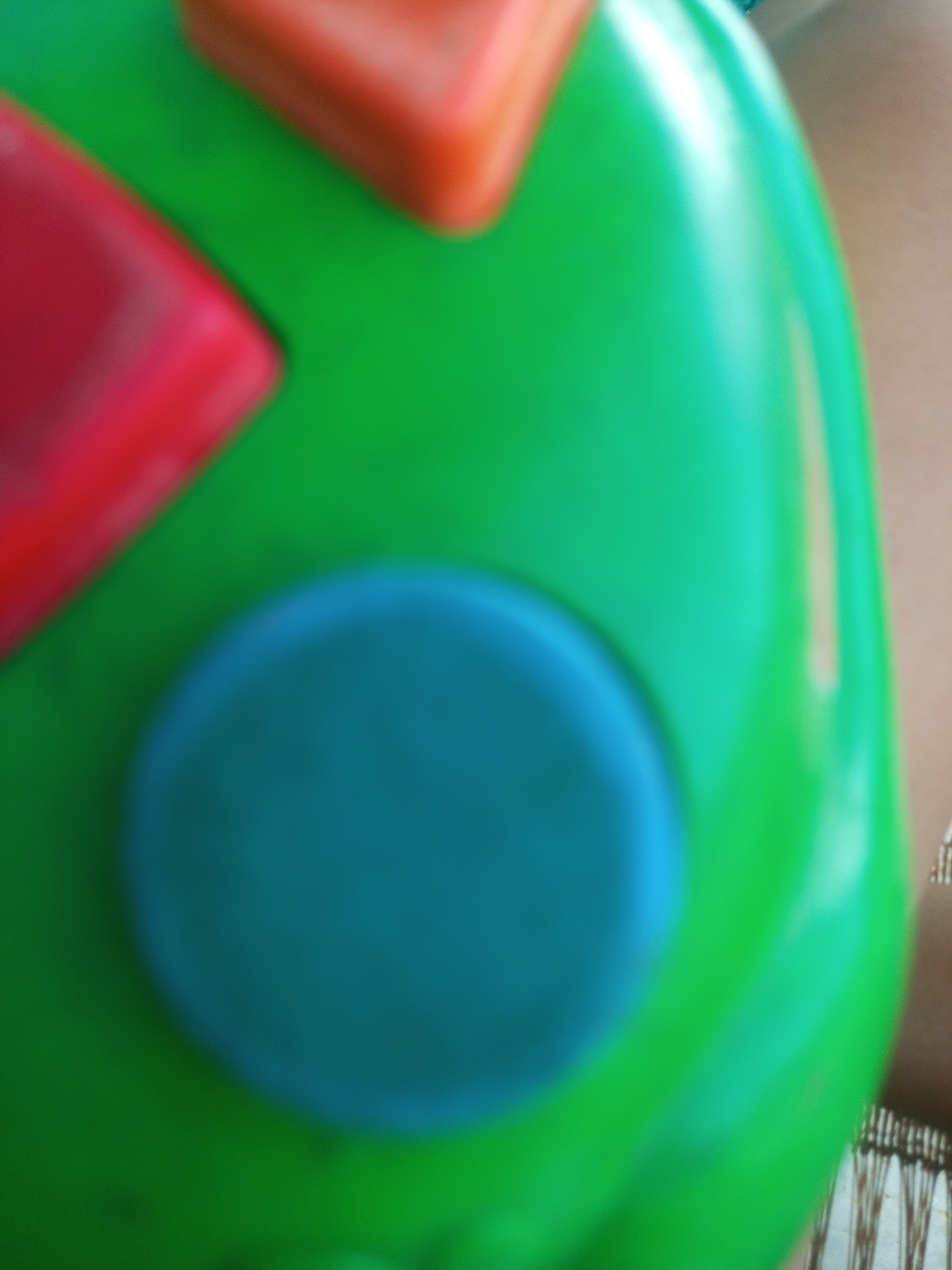 The image is a close-up, out-of-focus shot of a colorful plastic toy. The toy is primarily green, with a large, round, teal-colored button or dome. There are also red and orange shapes visible on the toy, which appear to be other buttons or decorative elements. The background is blurry and suggests a soft, neutral surface, possibly a table or floor, with some woven material visible on the right side. The image is not specific to any location in La Tagua, Venezuela, as it is an abstract close-up of a toy with no contextual environmental details.