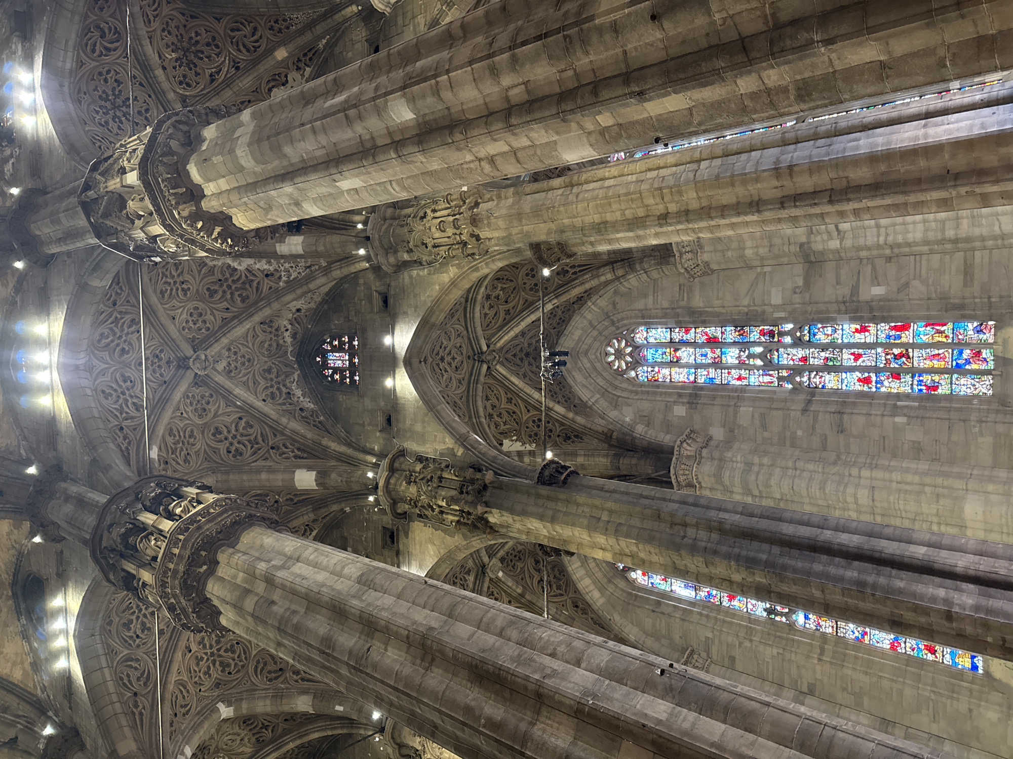 The image shows a low-angle view of the interior of the Duomo di Milano, also known as Milan Cathedral, in Milan, Italy. The cathedral's massive Gothic architecture is on full display, with towering stone columns supporting intricately carved vaulted ceilings. Sunlight streams in through tall, narrow stained-glass windows, casting colorful patterns on the stone surfaces. The stained glass windows depict various religious scenes and figures, typical of Gothic cathedrals. The lighting appears to be a combination of natural light from the windows and artificial lighting from fixtures mounted on the ceiling and walls, highlighting the architectural details. The scale of the columns and the ceiling suggests a vast and imposing interior space. There are no people visible in the image. The overall impression is one of grandeur, history, and religious artistry.