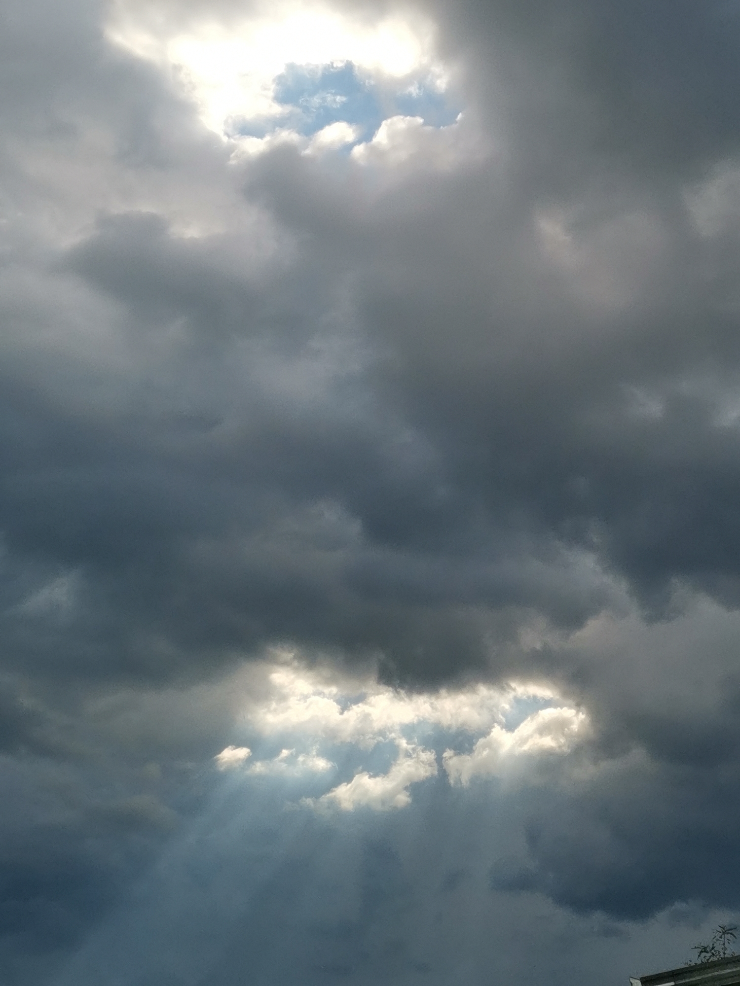 The image depicts a dramatic sky over Obligado, Paraguay, characterized by thick, dark storm clouds interspersed with patches of bright sunlight. In the upper portion of the frame, a large opening in the clouds reveals a patch of clear blue sky, with the sun shining through, creating a bright, almost ethereal glow. Below this, the clouds are heavy and gray, suggesting impending rain or a recent downpour. Further down, another, smaller break in the clouds allows sunlight to stream through in visible rays, illuminating some of the lower clouds and creating a dramatic contrast with the darker masses. The overall impression is one of dynamic weather conditions, with light and shadow playing a prominent role. The bottom edge of the image shows the hint of a structure, possibly a roof or building, with some vegetation visible to its right, indicating an outdoor scene in a populated area.