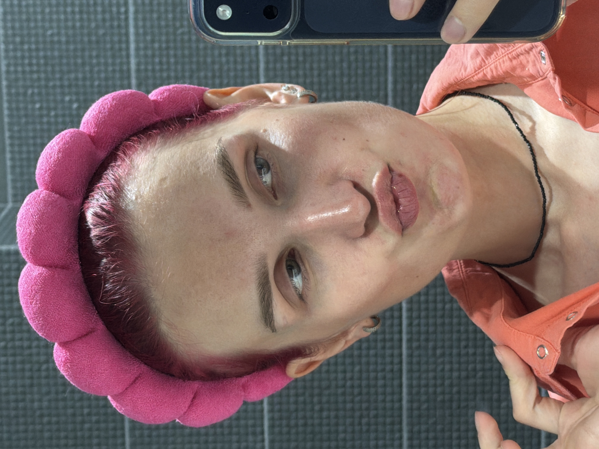 A close-up, vertically oriented selfie shows a person with fair skin, light-colored eyes, and pink-toned hair. They are wearing a bright pink, plush, padded headband, typically used during beauty routines. The person is dressed in a peach or coral-colored collared shirt, and a thin black beaded necklace is visible around their neck. A small hoop earring is also present in one ear.
The individual is looking directly into the camera with a neutral to slightly serious expression, their lips slightly parted and adorned with a light, muted pink lip product. The top portion of a dark-cased smartphone, including its camera lens, is partially visible at the top of the frame, indicating the person is holding the phone to take the selfie.
The background features a dark gray, textured surface, which could be a floor mat or wall. The lighting is bright and even, suggesting an indoor setting, likely during the daytime. The person's skin shows natural texture and appears somewhat moisturized, with some visible pores and minor marks. The pink hair at the hairline shows slight regrowth or darker roots. There is no visible text in the image.