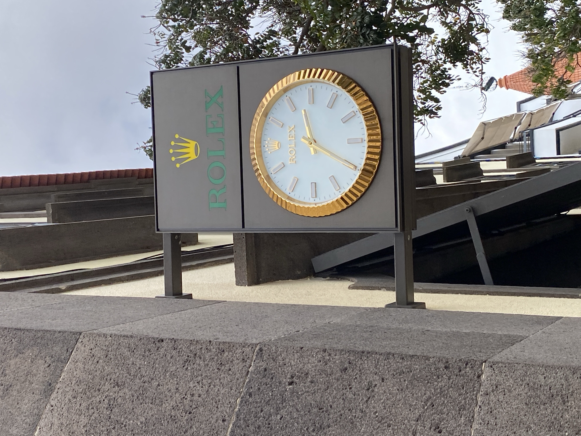The image shows a Rolex clock hanging on the side of a building in Funchal, Portugal. The clock is round with a gold-colored bezel and a light blue face. The Rolex logo, a golden crown, is visible above the word "ROLEX" in green letters. The building is made of grey stone, and there are trees with green leaves in the background. The sky is overcast.