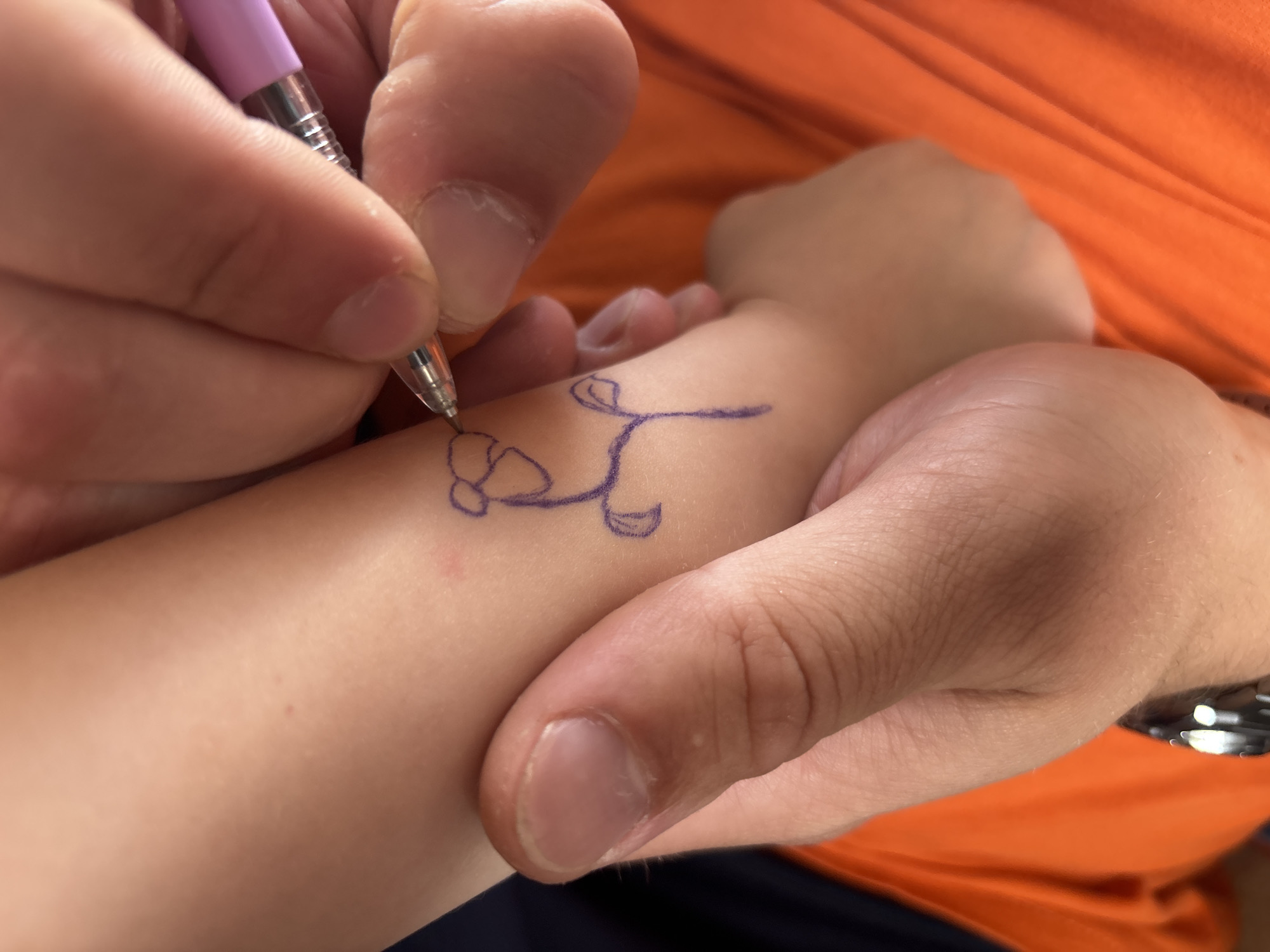 In the city of Niva, Russia, a close-up shot captures a person's hand drawing a flower on another person's arm with a purple pen. The drawing is simple and appears to be done with a marker. The arm is pale and smooth, and the person drawing is holding the pen with their index and middle fingers. The background is out of focus, but appears to be an orange fabric. The lighting is soft and natural.