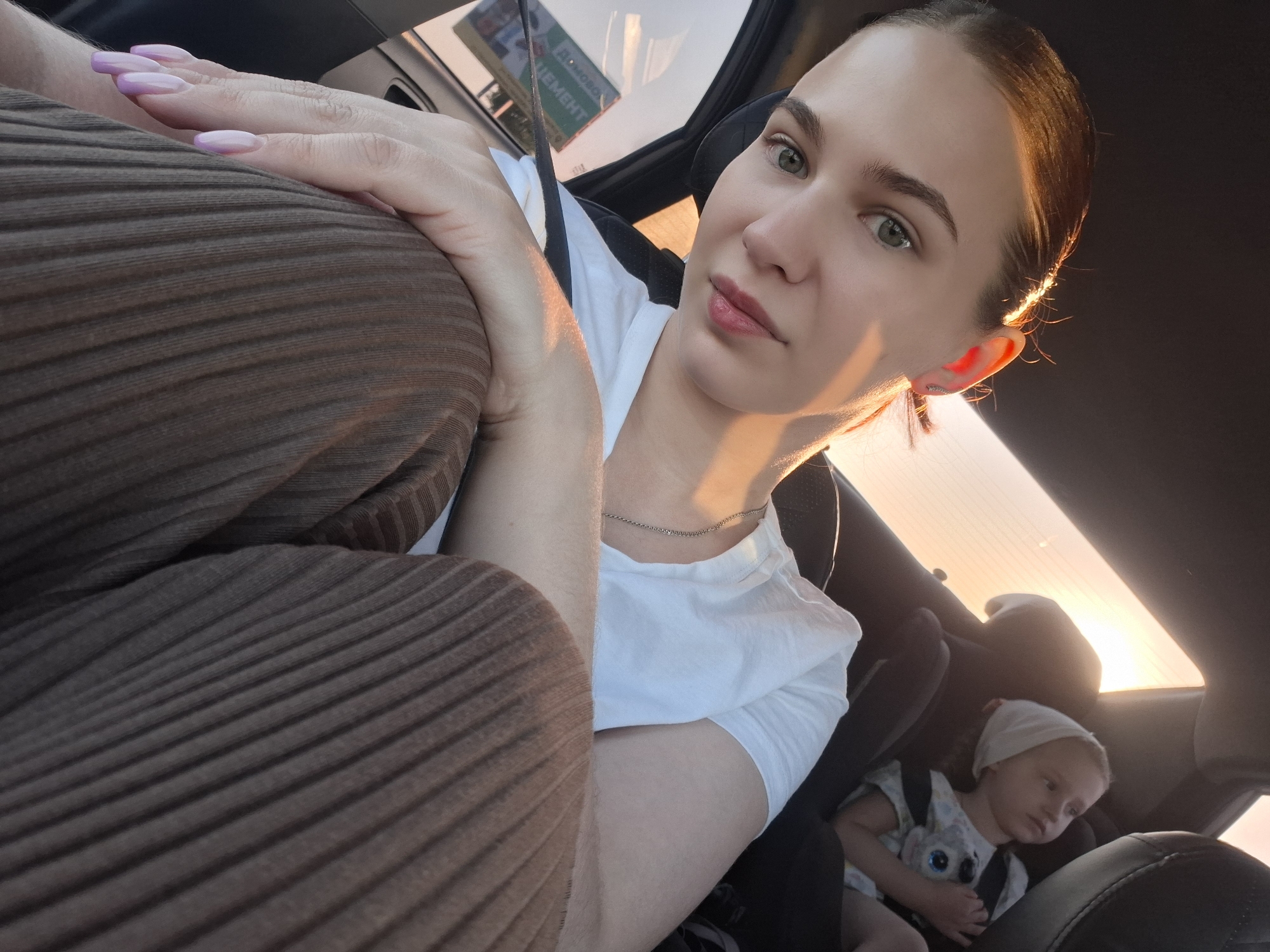 A woman is taking a selfie in the front seat of a car. She has brown hair pulled back, green eyes, and is wearing a white shirt and a silver necklace. Her hands are visible, with light purple nail polish. She is in the driver's seat with her left hand on what appears to be her lap, covered by ribbed brown clothing. Behind her, in a car seat, is a baby wearing a white head covering and a white and black shirt with a cartoon character on it. Outside the car window, the light suggests it is late afternoon or early evening. A sign is visible through the windshield. Based on the sign, the location is Гаревая, Russia.