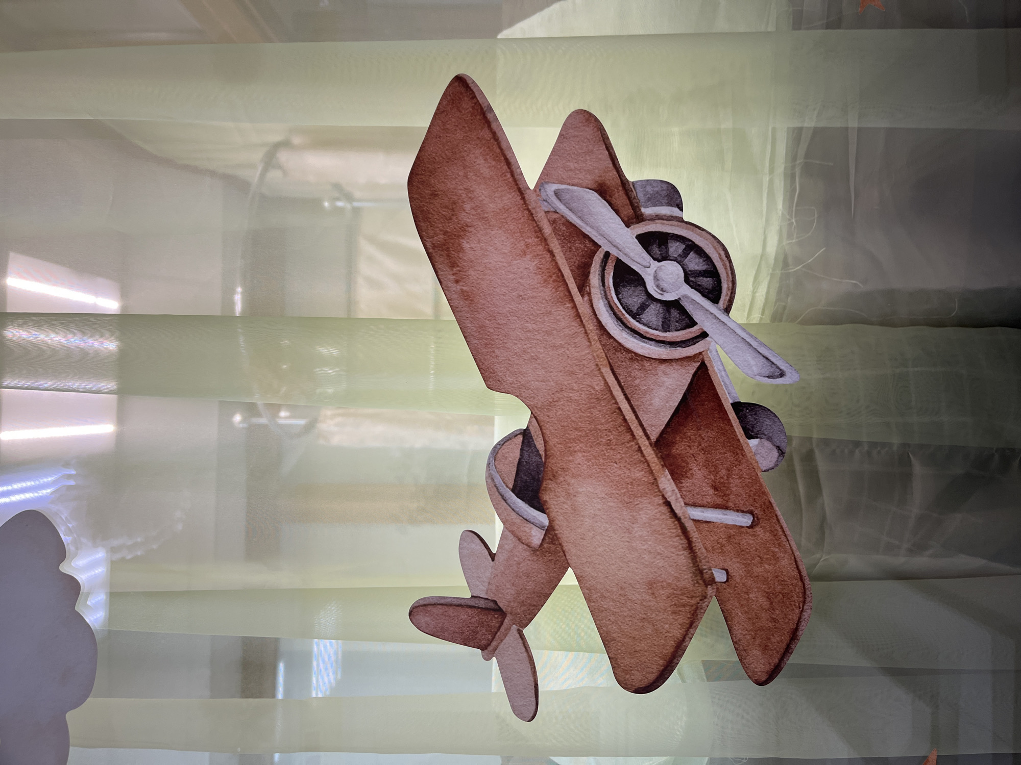 This media file features a watercolor-style illustration of a vintage biplane, depicted from a slightly elevated, angled perspective. The plane is predominantly brown with grey and white accents, showcasing a detailed propeller, engine, wings, and tail. It appears to be suspended or the illustration is placed against a backdrop of translucent, layered curtains. These curtains are a pale green color, with horizontal banding and subtle variations in opacity that create a soft, diffused light effect. The lighting suggests an indoor setting, possibly near a window due to the bright, horizontal light source visible on the left side of the frame, which also casts a silhouette of a cloud shape. The overall mood is whimsical and decorative, potentially intended for a child's room or a themed event. There is no text visible in the image. The location context of Chaikovsky, Russia is not directly indicated by visual cues within the image itself.