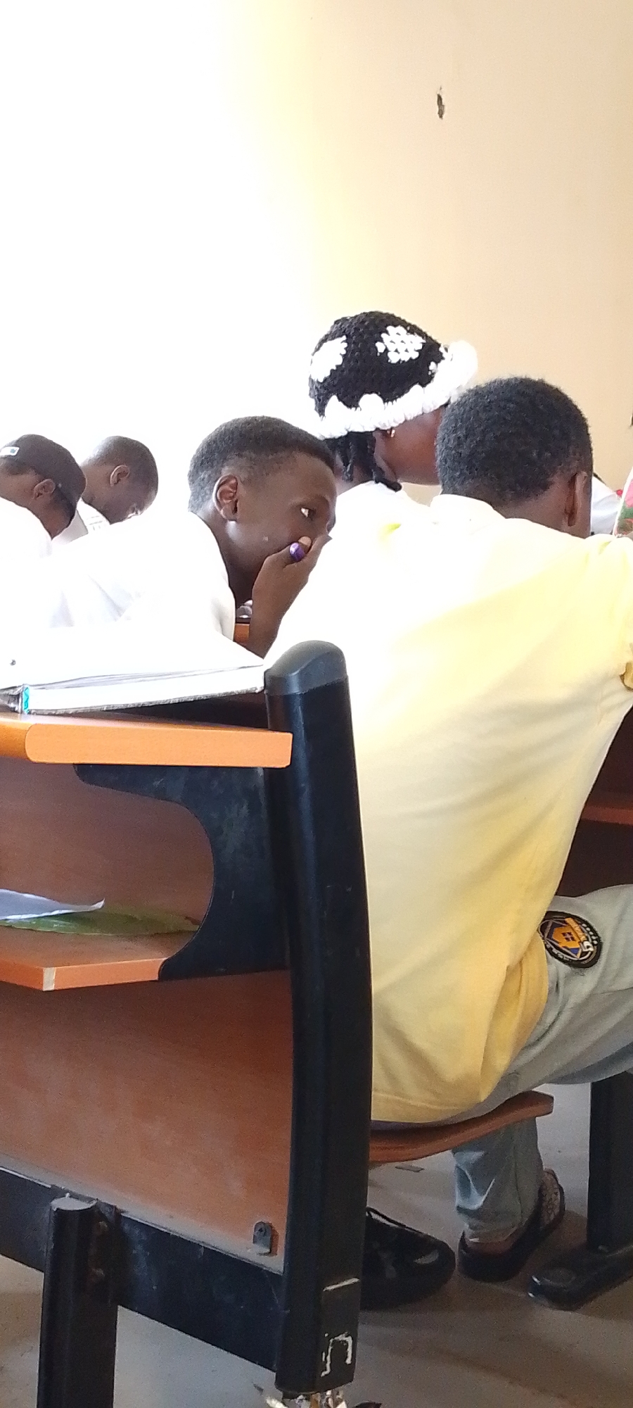 The image shows a classroom scene in Gumel, Nigeria, focusing on several young people seated at desks. In the foreground, a young person in a white shirt is looking to the side with their hand near their mouth, possibly in thought or speaking. To the left and further back, other individuals in white shirts are also seated at desks. A person in the background wears a black and white crocheted hat and a yellow shirt. The desks appear to be made of wood and metal. The overall environment suggests an educational setting.
