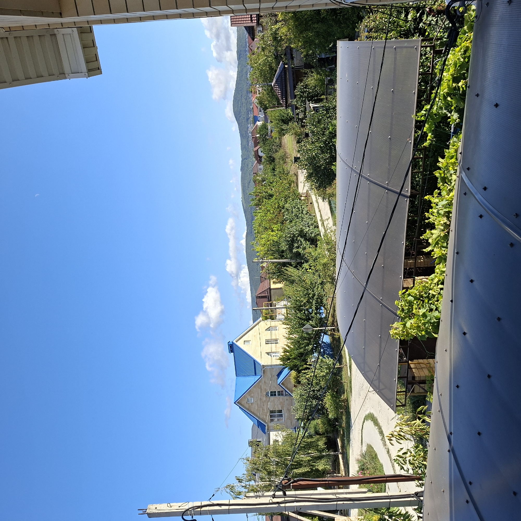 The image depicts a residential area under a bright blue sky with scattered clouds. In the foreground, the upper part of two structures with dark, translucent, ribbed roofs is visible. Beyond these, a curved paved path winds through a green landscape with various trees and bushes. A light-colored, two-story house with a prominent bright blue roof stands out among the greenery. Further in the background, a range of green, rolling hills forms the horizon, with more houses and trees dotting the landscape. A power pole with wires is visible on the left side of the frame. The scene is consistent with a suburban or semi-rural setting in Anapskaya, Russia, characterized by detached houses, vegetation, and a clear sky. No people are present in the image.