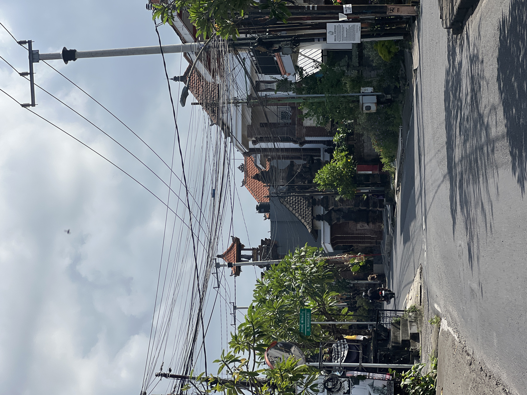 The image depicts a street scene in Denpasar, Indonesia, with a focus on a section of road winding through a residential or mixed-use area. The foreground is dominated by a gray asphalt road, with visible tire tracks and shadows cast by trees and utility poles. On the left side of the road, a sidewalk and some vegetation are visible, along with a street sign that reads "JL. NAGASARI" in green text on a white background, accompanied by text in an unfamiliar script above. A circular convex mirror is mounted on a pole near the sign, reflecting the scene.
Further into the image, a motorcycle with a single rider is seen moving away from the camera, heading down the road. The architecture of the buildings lining the street is characteristic of Balinese style, with pitched tiled roofs, ornate stonework, and traditional temple-like structures. Utility poles with a dense network of electrical wires crisscross the sky, a common sight in Indonesian urban environments. The sky is overcast with gray clouds, suggesting a possibly humid or impending weather event. Some trees with lush green foliage are interspersed among the buildings, adding a touch of nature to the urban landscape. Several signs, some with text and diagrams, are attached to utility poles and walls, indicating local regulations or advertisements. The overall impression is that of a typical, active street in a Southeast Asian city.