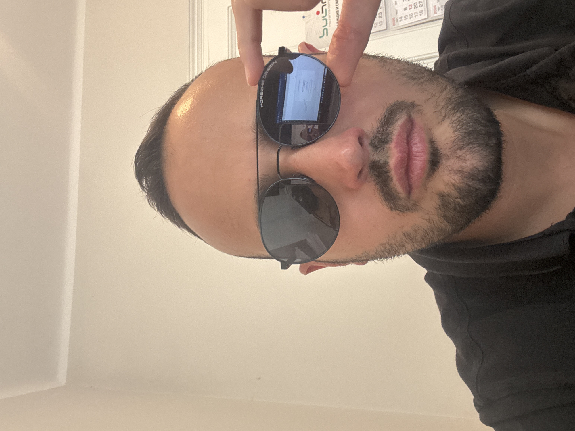 A man with dark hair and a beard is holding up a pair of round-framed sunglasses. His forehead is visible above the sunglasses. The reflection in the top lens shows a computer screen and a person. He is wearing a black shirt. The background includes a white wall and a calendar. Based on the logo "SuG" on the calendar, and "Млиция и сив" (Militia and gray) in Bulgarian, it's likely that the location is in Bulgaria, perhaps the city of Sofia.