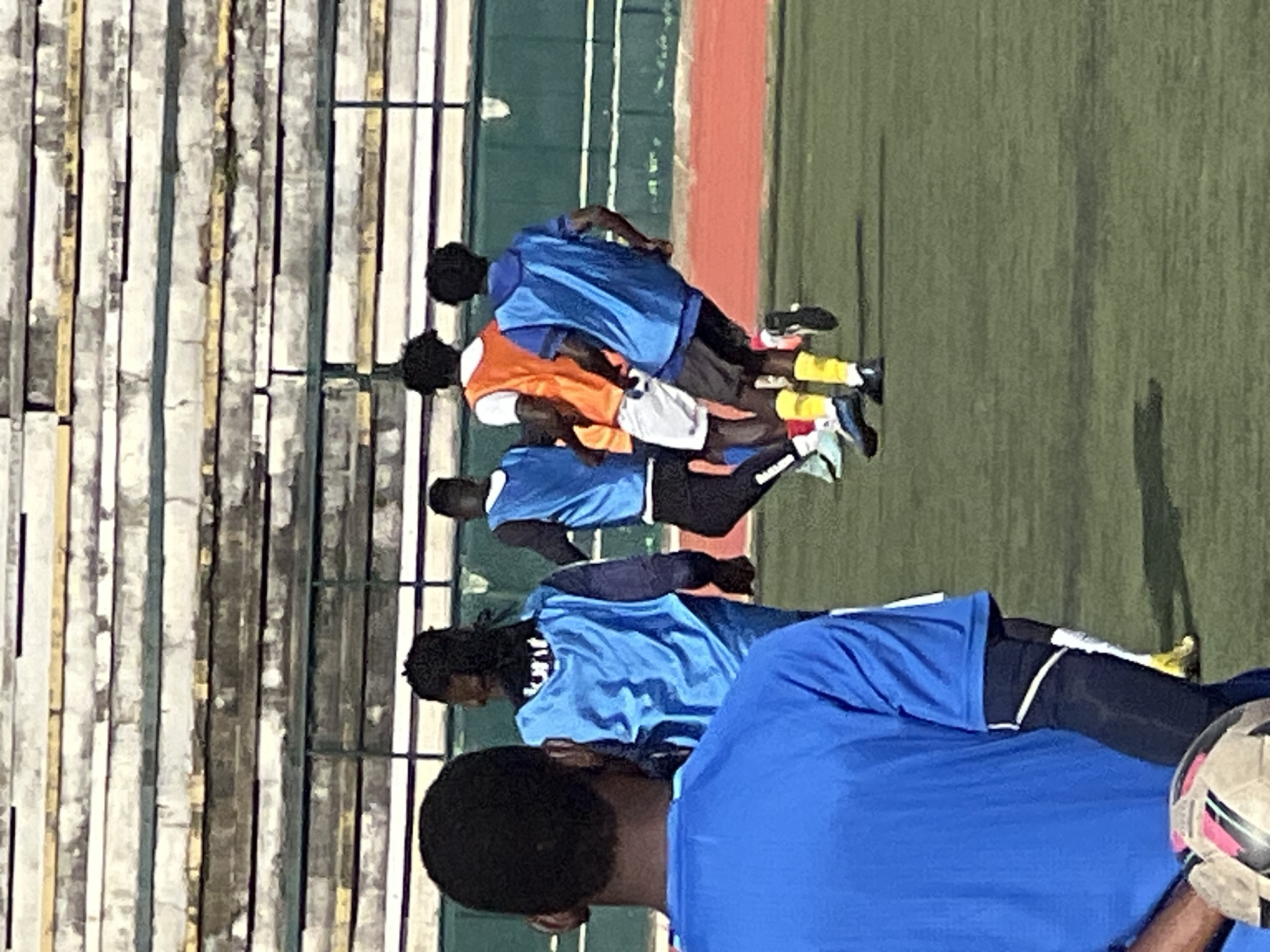 In the city of New Stadium, Nigeria, a group of young men in athletic wear are on a grass field, likely a soccer pitch. The foreground features a young man from the back, wearing a bright blue t-shirt and a black sleeve on his left arm, holding a soccer ball. In the middle ground, several other individuals are engaged in what appears to be a soccer training session. One person wears an orange vest over a white t-shirt and white shorts. Another individual is in a blue vest and dark shorts with bright yellow socks. The background shows stadium seating, with weathered concrete tiers and metal railings, suggesting a sports venue. The lighting indicates it is daytime, with shadows cast on the field.