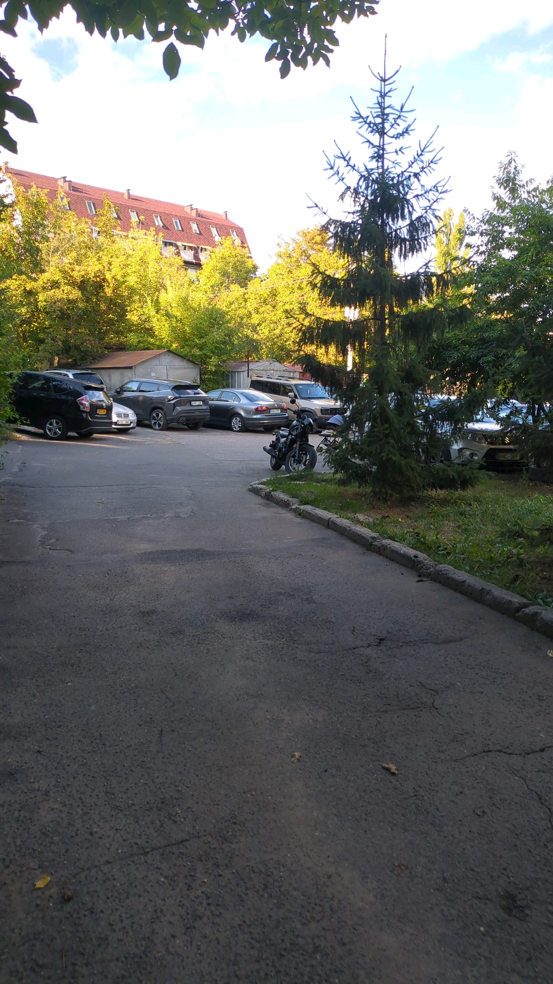 An outdoor, medium shot captures a paved parking lot adjacent to a tree-lined residential area in Chișinău, Moldova, under a bright, partially cloudy sky. In the background, a multi-story building with a red-tiled mansard roof is visible, surrounded by trees with yellow and green foliage, suggesting an autumn or late summer season. A variety of parked cars are present, including a black SUV, a silver sedan, and a light-colored sedan. A black motorcycle is parked on the right side of the lot, closer to the foreground. The pavement is cracked and worn, with some patches of grass growing along the edge. A large evergreen tree dominates the right side of the frame, casting shadows. The scene is quiet and appears to be unoccupied at the time of the photograph.