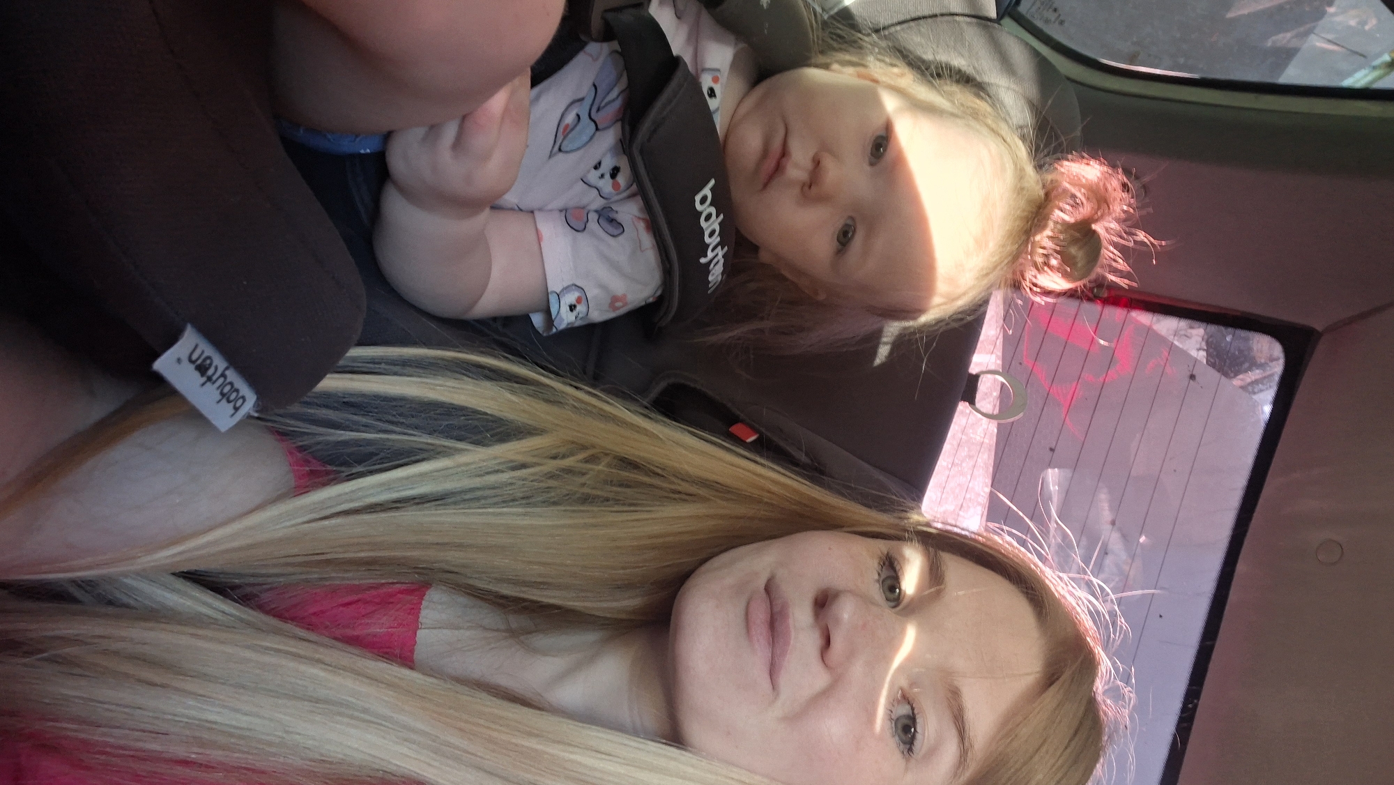This is a first-person perspective, close-up photograph taken from inside a car, likely in the city of Chaikovsky, Russia. A young child is seated in a car seat on the left, facing forward and looking towards the camera with a neutral expression. The child has blonde hair styled in a messy bun and is wearing a light-colored shirt with a cartoon bunny pattern. A woman, presumably the driver or passenger, is seated to the right, also looking towards the camera. She has long blonde hair and is wearing a bright pink top. Sunlight streams through the car windows, casting shadows and highlights on their faces. The interior of the car, including the car seat and window tinting, is visible. The overall scene suggests a casual moment during a car journey.