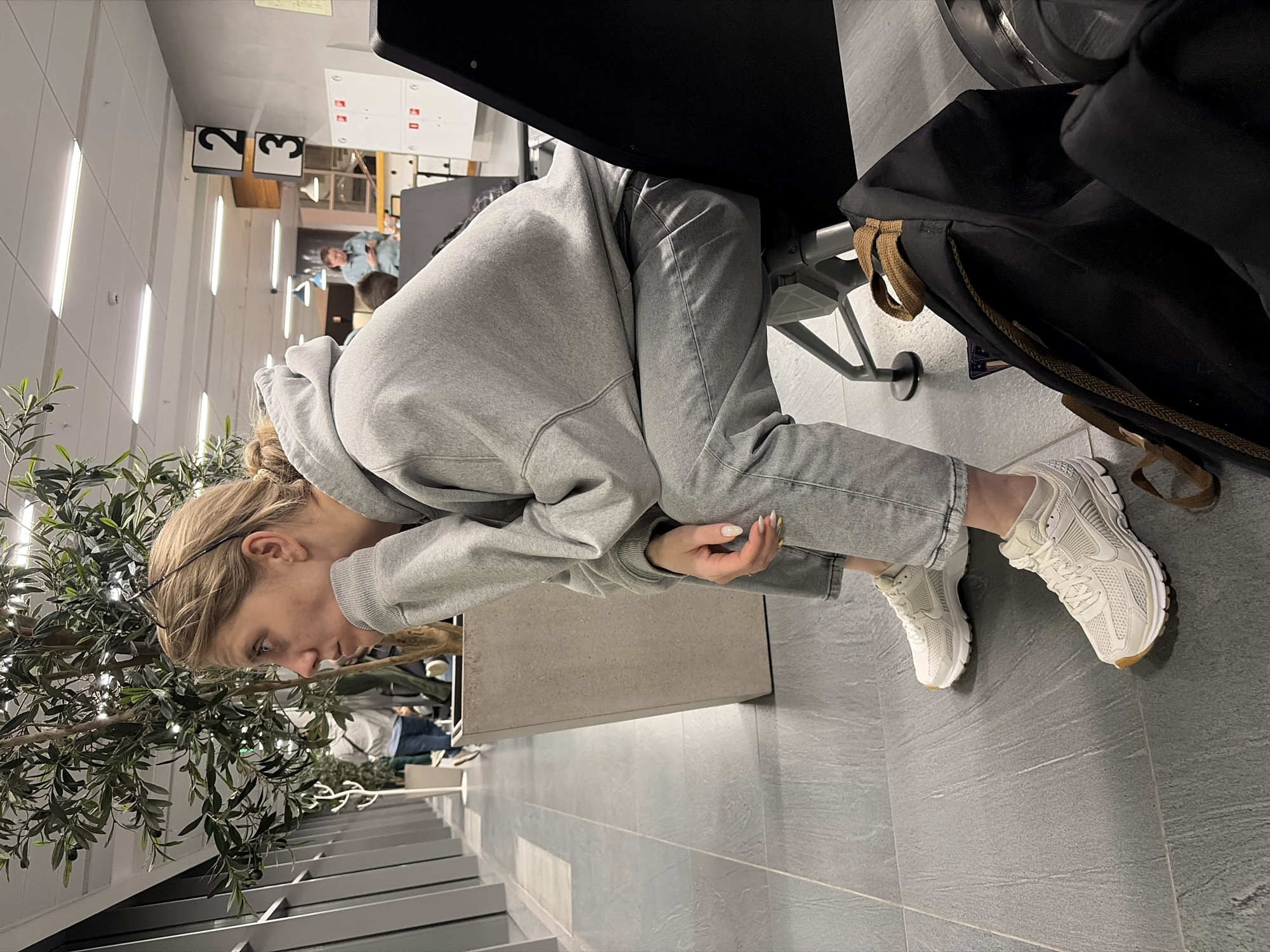 A young adult female is seated in profile, facing left, in what appears to be a public waiting area within the city of Zarechnaya (Gamovskoe S/P), Russia. She possesses light brown hair styled in a bun, with eyeglasses resting on her head. She wears a grey hooded sweatshirt, grey jeans, and white athletic shoes with brown soles. Her right hand supports her chin, and her left hand rests in her lap. To her immediate left is a tall artificial olive tree in a rectangular grey planter. The floor is composed of dark grey tiles. In the background, a white ceiling with fluorescent lighting is visible, along with black square signs displaying the numbers "2" and "3". Further back, a person in a light blue shirt is observed holding a mobile phone. A white wall cabinet with red square elements is present on the far right. A black backpack with brown straps and a portion of dark luggage rest on the floor next to the seated female.