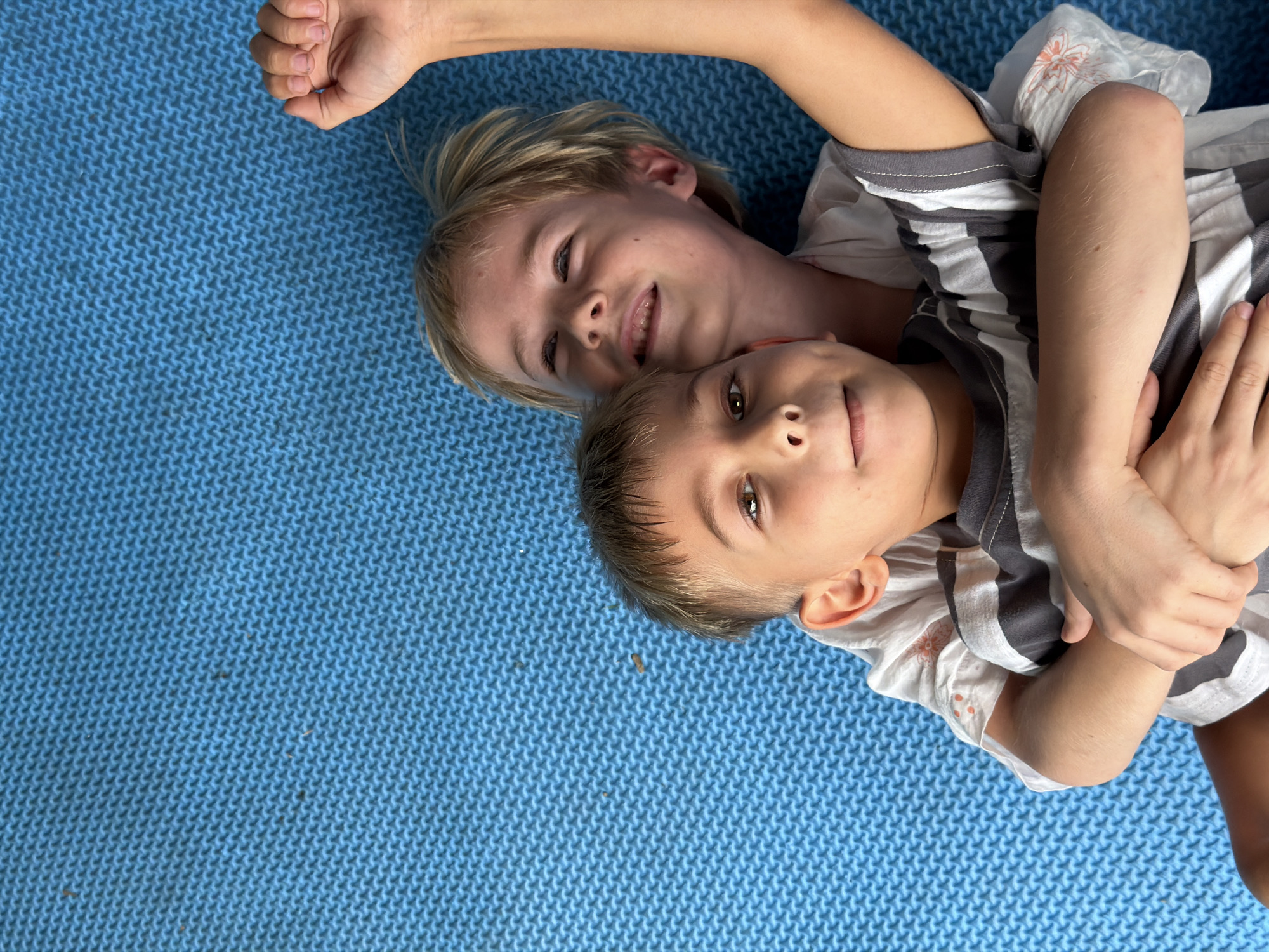 Two children lie on a blue textured surface. One child, with blonde hair, is positioned above the other, who has shorter brown hair. The blonde child's arm is extended above their head. Both children are wearing striped shirts. The location is unspecified, not Kuta, Indonesia.