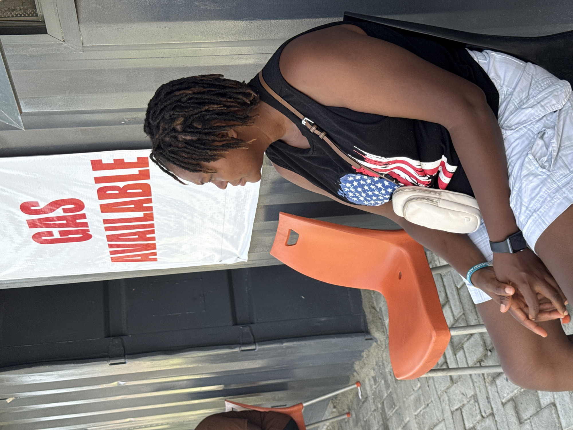 A woman sits in an orange plastic chair in front of a building with a sign that reads "Gas Available" in Ado, Nigeria. She wears a black tank top with an American flag design, light-colored shorts, and a smartwatch. A small, light-colored purse hangs from her shoulder. Her hair is styled in dreadlocks. Her hands are clasped together. The ground is paved with interlocking concrete pavers.