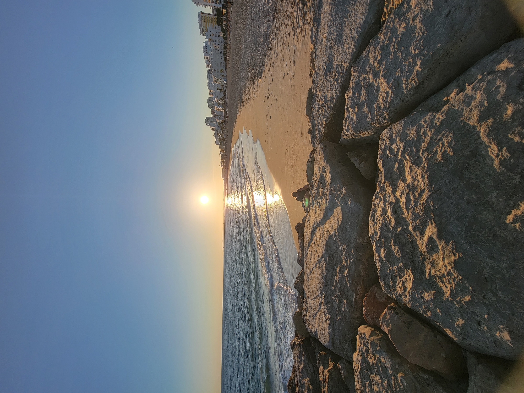 This is a long shot, wide-angle photograph of a beach at Quarteira, Portugal, at sunset. The sun is visible in the upper right quadrant of the image, casting a warm glow across the scene. The ocean is on the left, with gentle waves rolling onto the sandy shore. In the foreground, large rocks are visible, some of which are partially submerged in the water. Along the beach, a row of white buildings forms a skyline. The sand is mostly empty, with a few people scattered along the shore. The sky is clear and blue, with a gradient towards a lighter shade near the horizon.