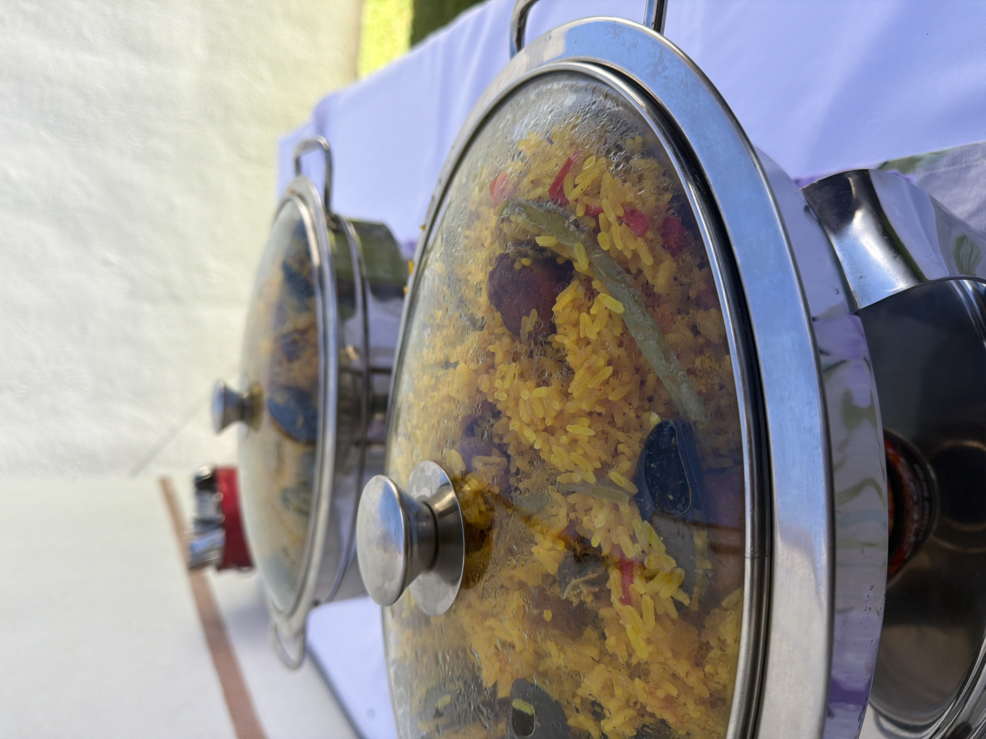 The image shows two chafing dishes with glass lids and metal frames. The dishes appear to contain a yellow rice dish with various vegetables, possibly paella or a similar rice-based meal. They are set on a white surface, likely a table or serving area, with a white tablecloth in the background. The scene appears to be at an outdoor event or buffet. The location is Unknown, Unknown.