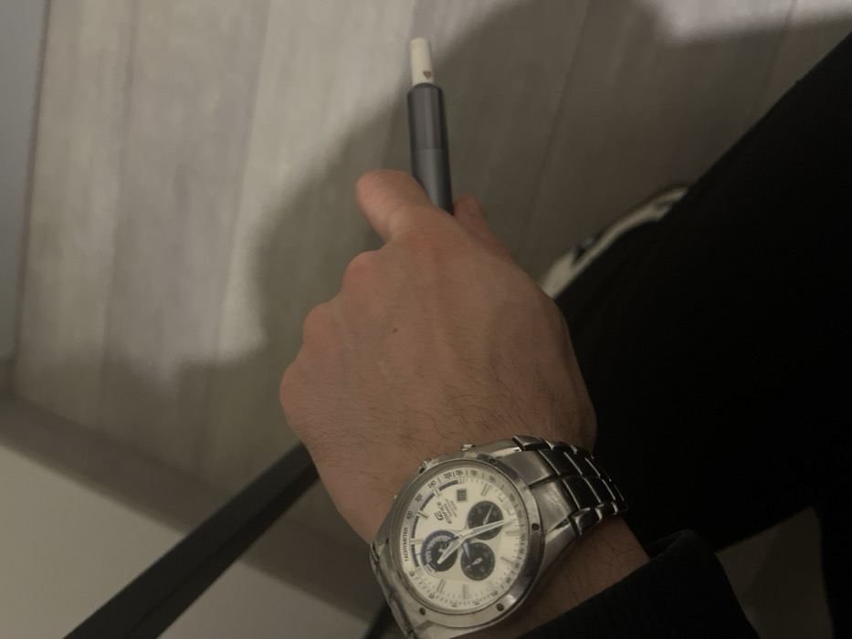 The image displays a close-up view of a left arm and hand in an indoor setting within Sallanches, France. A light-skinned individual wears a silver-toned Casio Edifice chronograph watch, featuring a white dial with three sub-dials, a tachymeter scale, and a metal link bracelet, on the left wrist. The inscriptions "EDIFICE" and "CHRONOGRAPH" are visible on the watch face. The hand grasps a dark grey cylindrical device, identified as an IQOS heated tobacco product, from which a white HEETS stick with a distinct red logo is inserted and protrudes. Portions of black apparel are visible in the lower left section of the frame. The background shows a light-toned, horizontally planked floor or surface, along with a dark, vertical structural element, likely part of furniture, positioned on the far left.