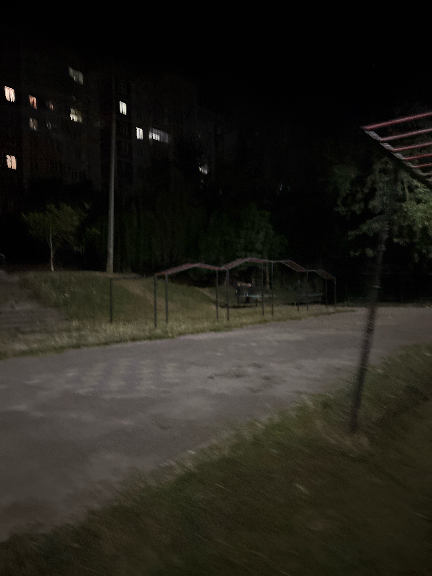 The image captures a dark, possibly nighttime scene in Ternopil, Ukraine. In the background, a multi-story residential building with illuminated windows is visible. In the foreground, there is a paved area, likely a pathway or courtyard, adjacent to a grassy slope. Several metal structures, possibly part of a playground or park furniture, are present on the slope, some with red-colored tops. Trees and foliage surround the area, adding to the somber atmosphere. The scene is devoid of any people or discernible activity, presenting a static, somewhat deserted environment.