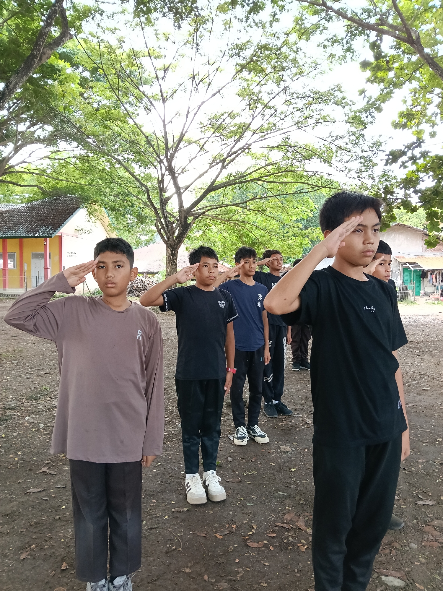A group of seven young men are standing in a line on an outdoor dirt surface, saluting with their right hands to their foreheads. The young men are wearing casual clothing, including t-shirts and pants in black, brown, and blue, and some are wearing sneakers. Behind the line of young men, there are trees and a partial view of a low, yellow building with a gray roof. The setting appears to be in a residential or institutional area, possibly in Lhokseumawe, Indonesia, given the prompt's location specificity. The lighting is overcast, casting soft shadows on the ground. The ground is covered with dirt and scattered leaves.
