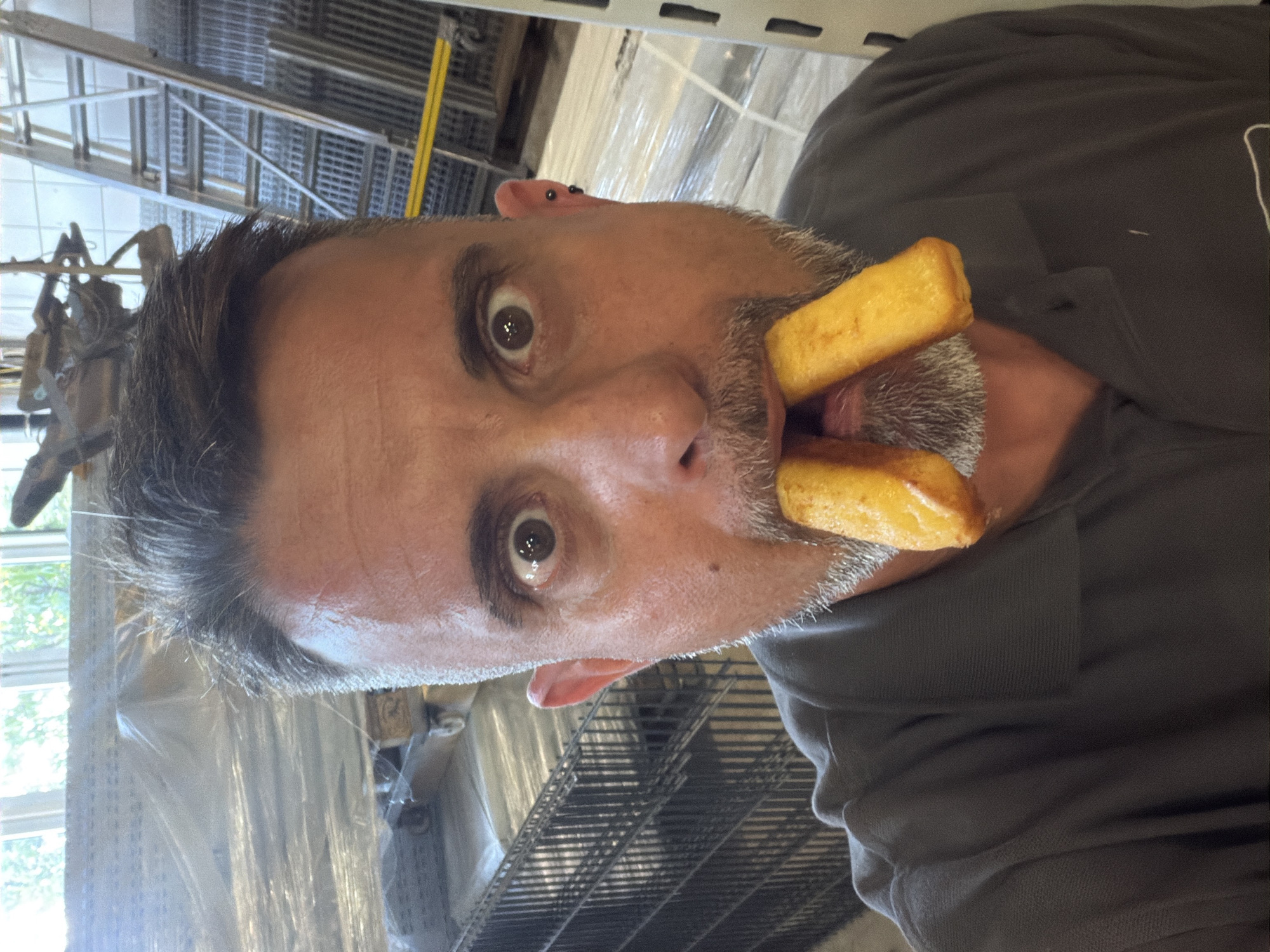 A close-up, front-facing selfie of a man with a grey beard and short, dark hair, who is in a warehouse in Leudelange, Luxembourg. He is wearing a grey polo shirt and has two french fries in his mouth. His eyes are wide open, and he has a surprised expression. The background shows stacks of items wrapped in plastic, metal shelving units, and a ladder.