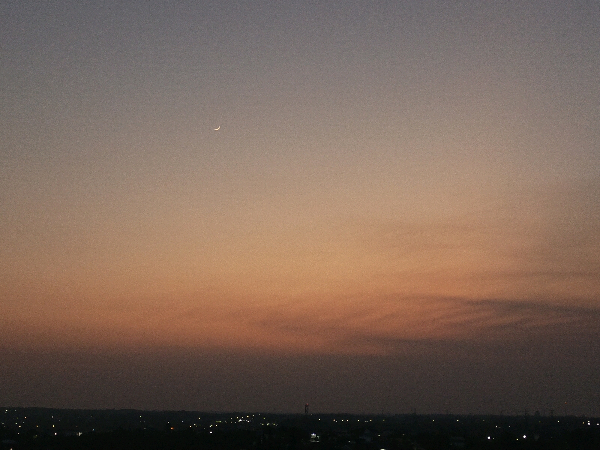 The image shows a sunset sky over the city of Banjarbaru, Indonesia. The sky is a gradient of colors, from a pale grey at the top to a deep orange and then to a dark purplish-grey near the horizon. A small, bright crescent moon is visible in the upper left quadrant of the sky. Below the sky, a dark silhouette of the city is visible, dotted with small, bright lights, indicating buildings and streetlights. In the middle distance, a tall tower with a red light at its top is present. The overall impression is one of twilight settling over an urban landscape.