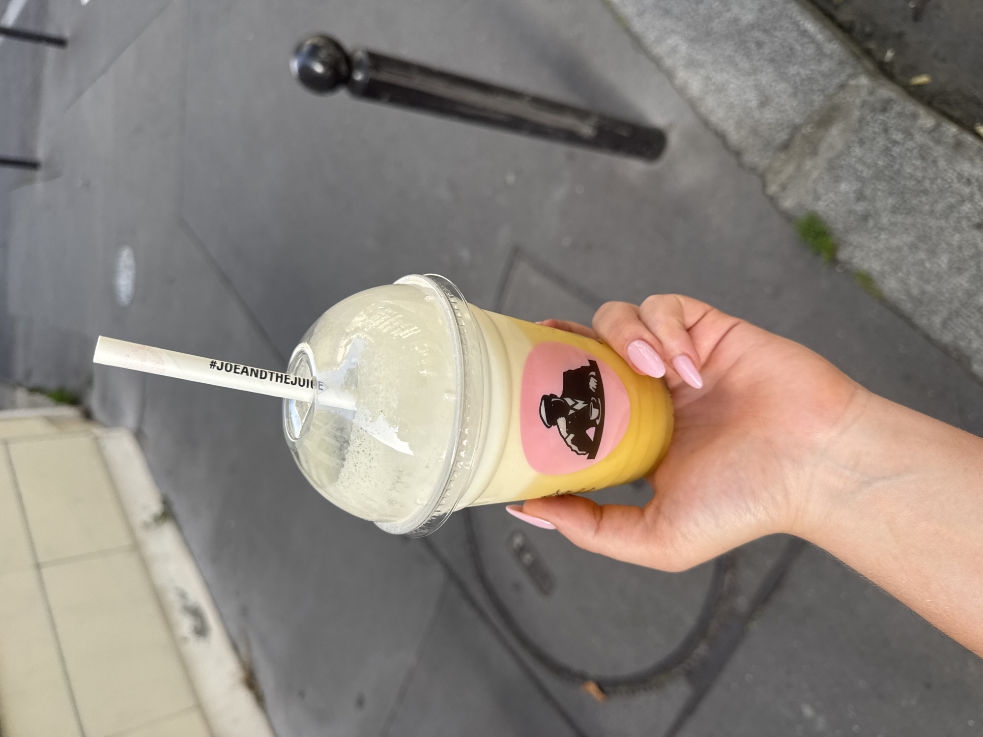 A hand holds a yellow and white juice drink from "Joe and The Juice" on a sidewalk in Paris, France. The drink has a clear domed lid, a paper straw with "#JOEANDTHEJUICE" printed on it, and a pink label with a black graphic. The hand has light skin and pale pink nails. The sidewalk is gray with darker gray patches and a dark metal post is visible in the background. There is some greenery growing on the edge of the sidewalk.