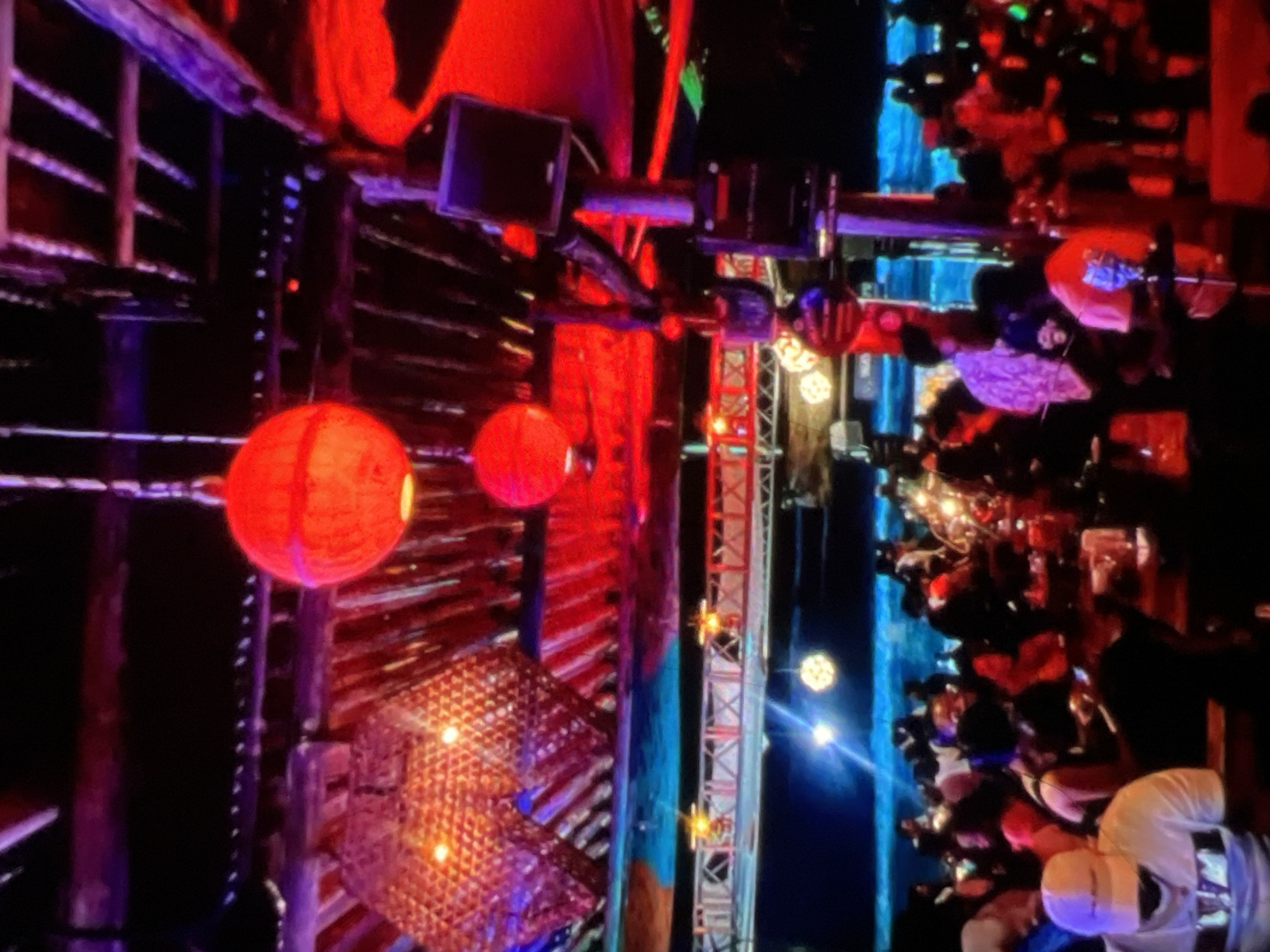 The image depicts an indoor scene at night, likely a nightclub or bar in Lagos, Nigeria, characterized by vibrant red and blue lighting and a crowded atmosphere. Large, spherical red lanterns are suspended from the ceiling, contributing to the ambient lighting, along with what appears to be a textured, possibly bamboo-like, decorative element on the left. The ceiling structure is visible, with visible beams and corrugated metal, suggesting a rustic or industrial aesthetic.
Below, a densely populated area shows patrons seated at tables and standing, engaged in social activities. Several individuals are visible, with varying attire, including a person in the foreground wearing a white cap and t-shirt. The environment appears to be a lively social gathering. The presence of speakers mounted on a pole and the overall ambiance suggest a venue for entertainment and socializing. The blue and red lighting creates a dynamic and energetic mood within the space.
