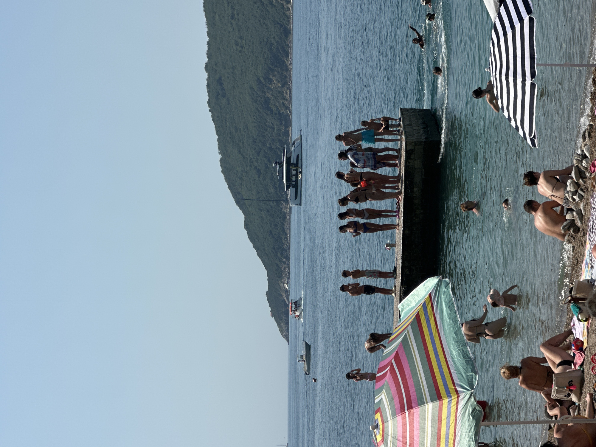 The image depicts a sunny day at a beach in Budva, Montenegro. The scene is populated with numerous people enjoying the water and the shore. Several individuals are standing on a concrete pier, some appearing to be preparing to dive into the blue sea. Others are swimming or wading in the water. A large yacht is visible in the background, anchored near the shore. Two beach umbrellas, one with black and white stripes and another with colorful stripes, provide shade for beachgoers. The overall atmosphere suggests a leisurely summer day with clear skies and calm waters. The foreground shows a rocky shoreline with towels and bags. The time of day appears to be afternoon, given the bright sunlight casting shadows.
