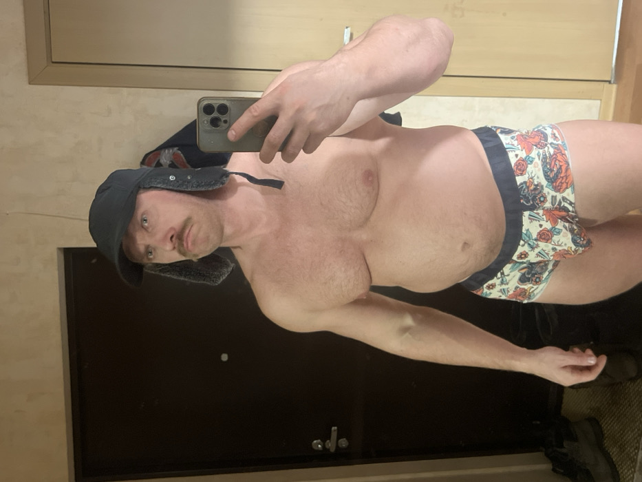 A man is depicted in a mirror selfie, shirtless and wearing patterned briefs and a trapper hat. The image is rotated 90 degrees clockwise.
The man, with fair skin and light brown hair, is looking directly at the camera with a neutral expression. He has a distinct mustache and appears to have a muscular build, particularly in his arms, while also showing some body fat around his abdomen. He is holding a dark-colored smartphone, likely an iPhone with multiple camera lenses, in his right hand to take the photo, which is reflected in the mirror.
He is wearing a dark blue or black trapper hat with ear flaps that are tied up. Below his waist, he wears white briefs with an intricate pattern featuring what appear to be old-school tattoo designs, including skulls, roses, anchors, and other nautical or traditional motifs, against a light background. The briefs have a dark band around the waist and leg openings.
The background visible in the mirror's reflection shows an indoor setting. To the left, there's a dark brown or black door, possibly with a handle or knob faintly visible. Above the door, a light-colored wall or trim can be seen. On the right side of the frame, a lighter-colored floor or surface is visible, and some dark objects, possibly shoes, are on the floor. No text is visible in the image.