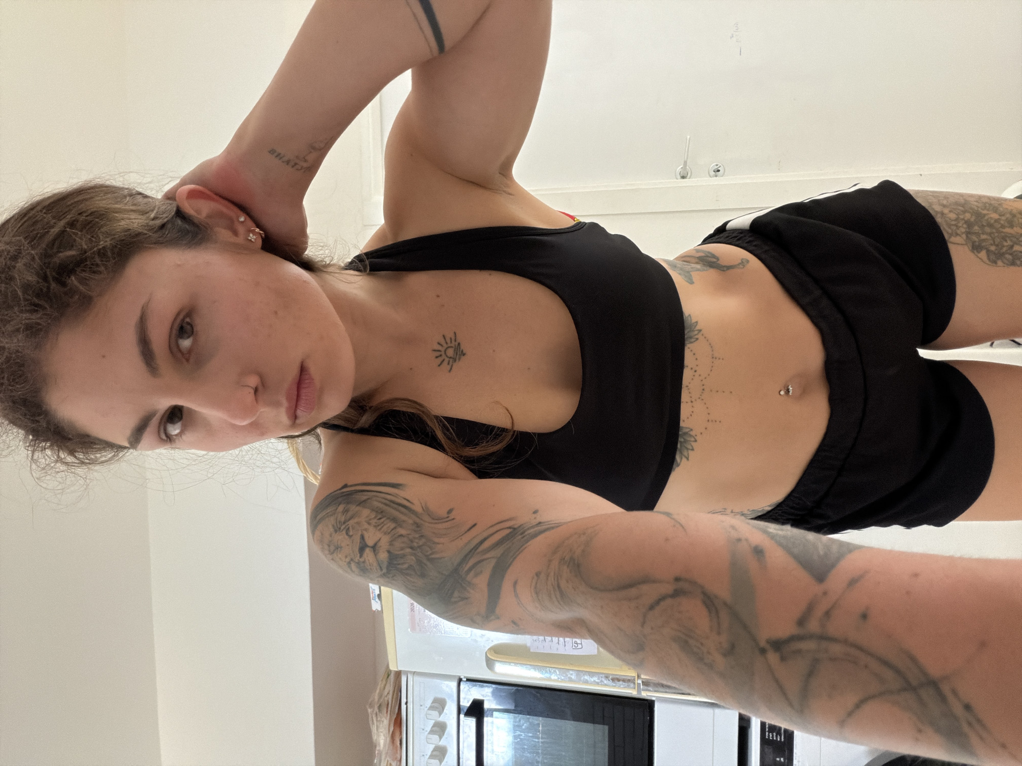 A woman is posing indoors for a selfie-style photograph. She has light skin, brown hair pulled back, and visible tattoos on her arms, torso, and potentially her leg. She is wearing a black sports bra and black shorts with white stripes. Her right arm is raised with her hand behind her head, while her left arm is extended towards the camera. A kitchen area is visible in the background, including a white oven with a black door and white cabinets. The location is Kiryat Bialik, Israel.