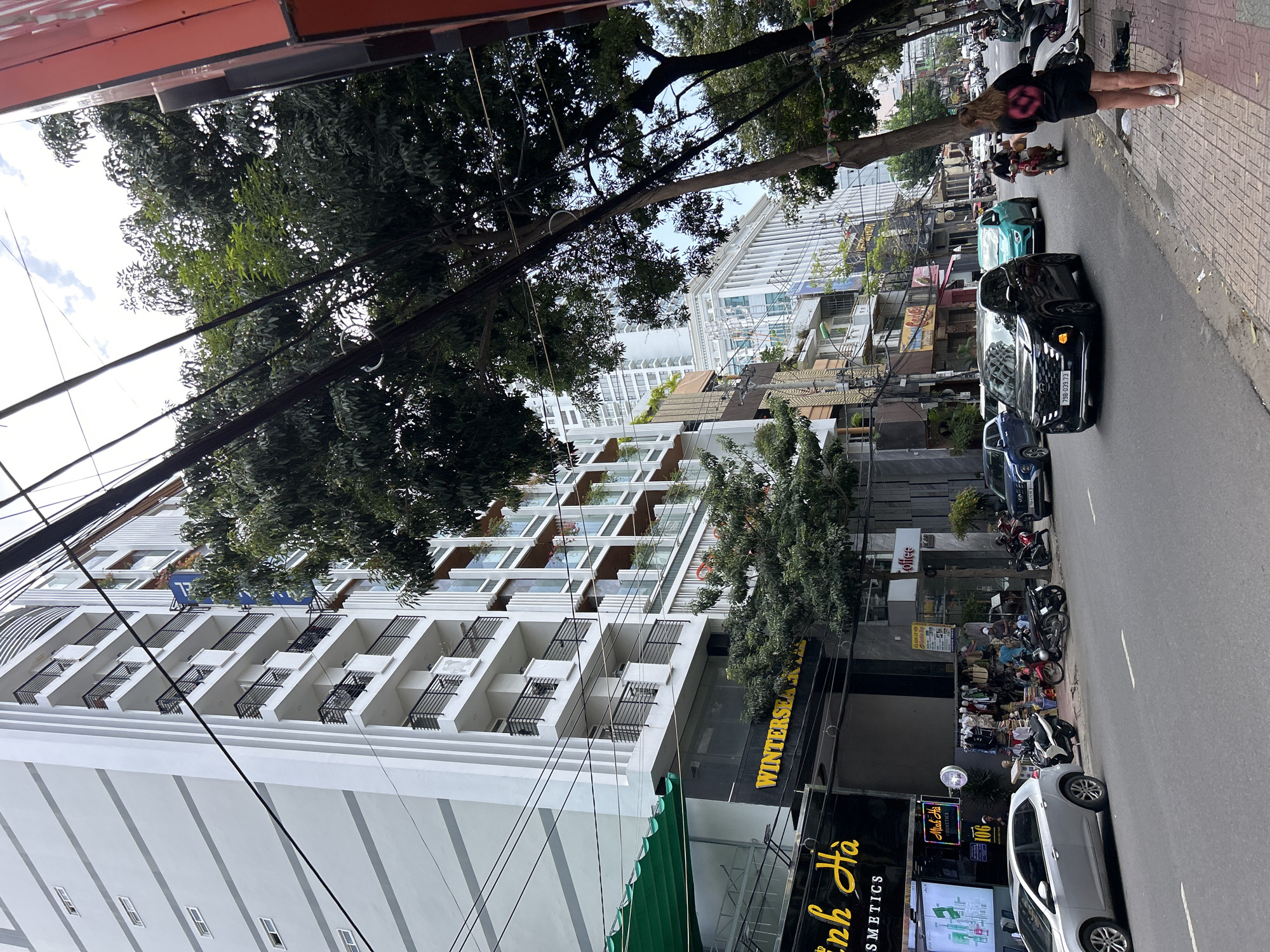 This street scene, captured during daytime in Nha Trang Ward, Vietnam, depicts a busy urban environment. The image, oriented vertically, shows a street running horizontally across the midground, flanked by buildings and trees, under a bright sky.
On the left side of the street, a tall, modern white building with multiple floors and numerous balconies dominates the frame, suggesting an apartment complex or hotel. Below this building, at street level, various storefronts are visible. One prominent sign displays "WINTERSEA HOTEL" in yellow lettering, while another nearby shop sign reads "Minh Hà COSMETICS" alongside Vietnamese text "Mỹ phẩm Minh Hà" and a house number "106". Further down the street, another sign for "COFFEE" can be seen. Overhead, a dense network of black electrical or communication wires crisscrosses the sky, partially obscured by lush green trees that line the thoroughfare.
The right side of the street features a paved sidewalk where a person with long blonde hair, wearing a black top with a distinctive pink/red circular design on the back, walks away from the camera. The street itself is lined with parked vehicles, including several cars and numerous motorbikes. A black Hyundai sedan is prominent in the foreground, displaying its license plate "79D-039.73." Behind it, a light blue car and other vehicles are parked. In the distance, more buildings and urban infrastructure are visible under a partly cloudy sky. The overall scene conveys the bustling atmosphere of a city street in Vietnam.