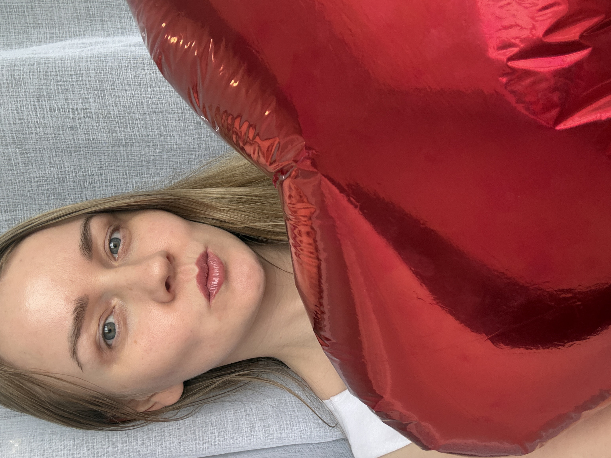 A young woman with fair skin and light brown hair is lying on a gray textured surface, possibly a sofa or bed. She faces the camera with a neutral expression. Her eyes are light-colored, and she has full lips with a shiny finish. A large, inflated red heart-shaped balloon partially obscures her face and upper body, filling a significant portion of the frame. The scene appears to be indoors. The image was taken in a residential area of the city of Chaikovsky, Russia.