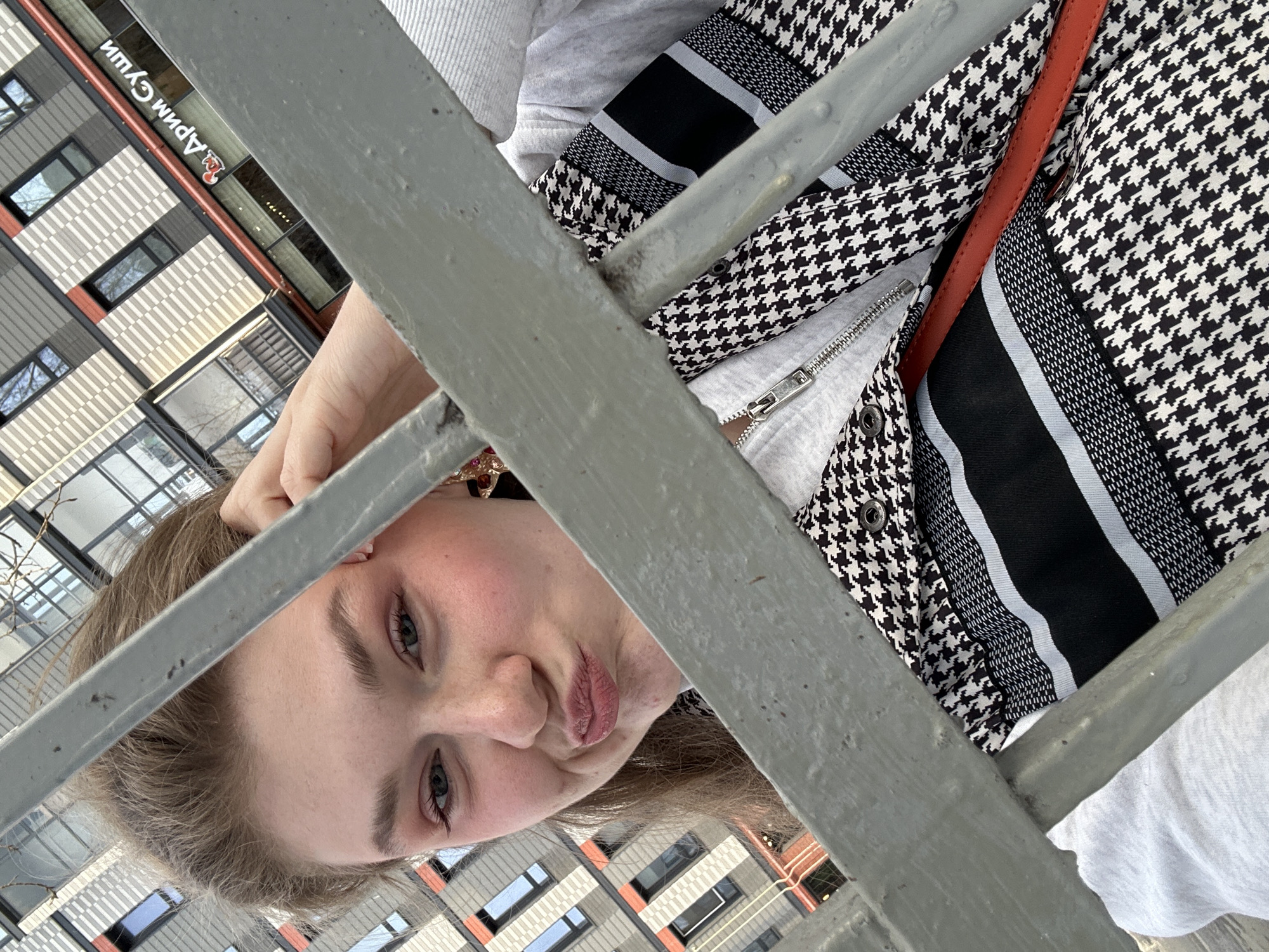 A young adult female is depicted in a close-up, angled shot, peering directly at the camera through a gray metal barrier composed of horizontal bars. Her head is tilted, and her lips are pursed in a playful expression. Her left hand is raised, with fingers touching the hair near her right temple. She wears a light grey zippered garment underneath a black and white houndstooth patterned jacket or vest, which also features horizontal black bands with thin white stripes. A red strap, likely from a bag, is visible across her chest. In the background, a multi-story modern building with a distinctive facade is visible. The facade consists of alternating light-colored and dark grey rectangular panels, with some reddish-orange accents around windows and as a horizontal band. A prominent sign on the building displays white Cyrillic text "Дрим Суши" (Dream Sushi) accompanied by a red, stylized seafood-related logo. This "Dream Sushi" establishment is located in the city of Kazan, Russia, consistent with branches found in modern complexes, such as those on Chistopolskaya Street. The scene is illuminated by natural light, and reflections of bare tree branches are visible in the building's windows.