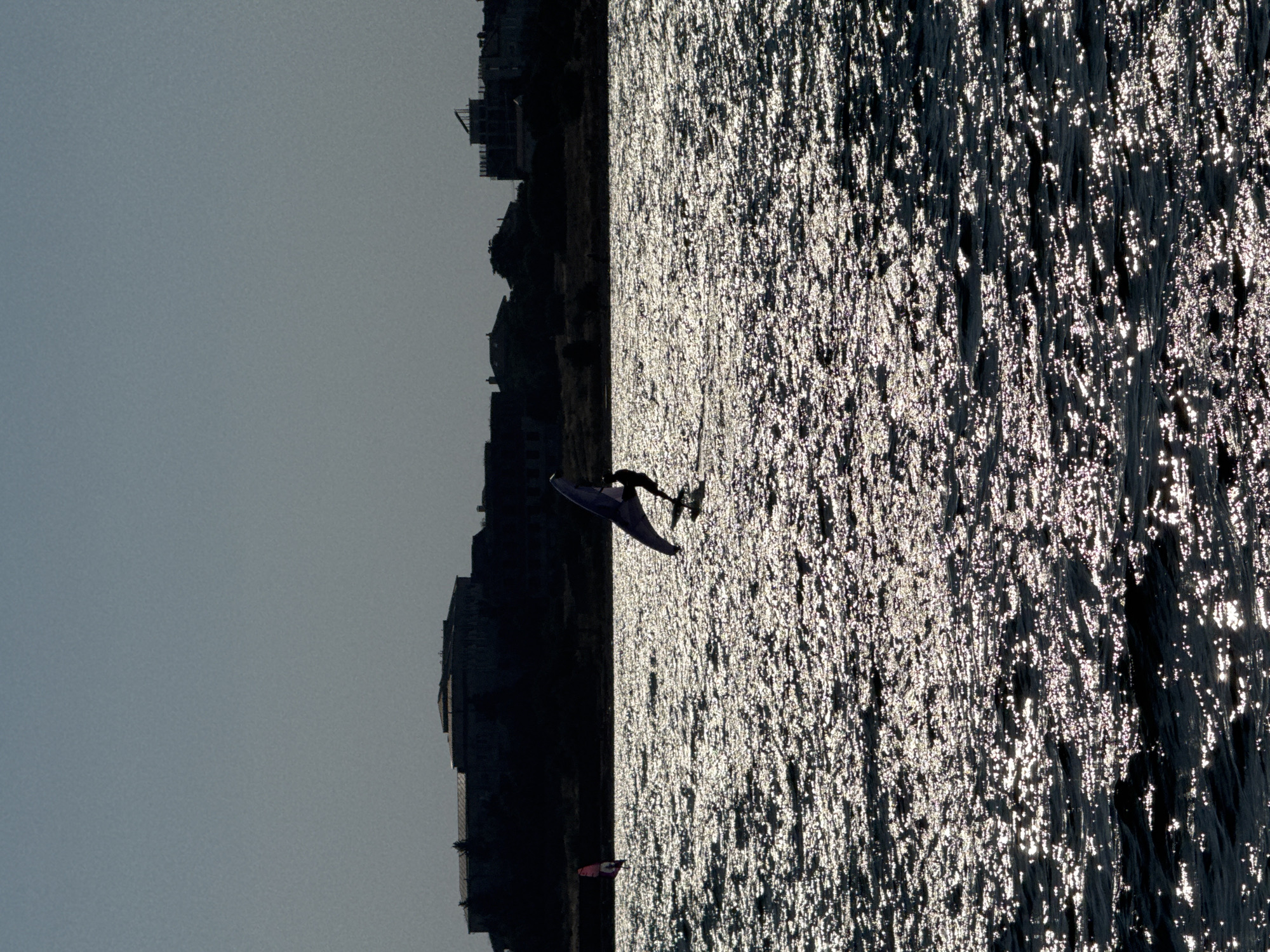 Two windsurfers are on the water. The windsurfer in the foreground is on the right side of the image and is silhouetted against the bright water. The windsurfer is wearing dark clothing and is holding a sail. The windsurfer in the background is on the left side of the image and is also silhouetted. The windsurfer is wearing dark clothing and is holding a sail. The water is choppy and the sun is glinting off the waves. In the background, there is a dark silhouette of buildings on a hill. This appears to be a view of the coast of Sevastopol, Ukraine.