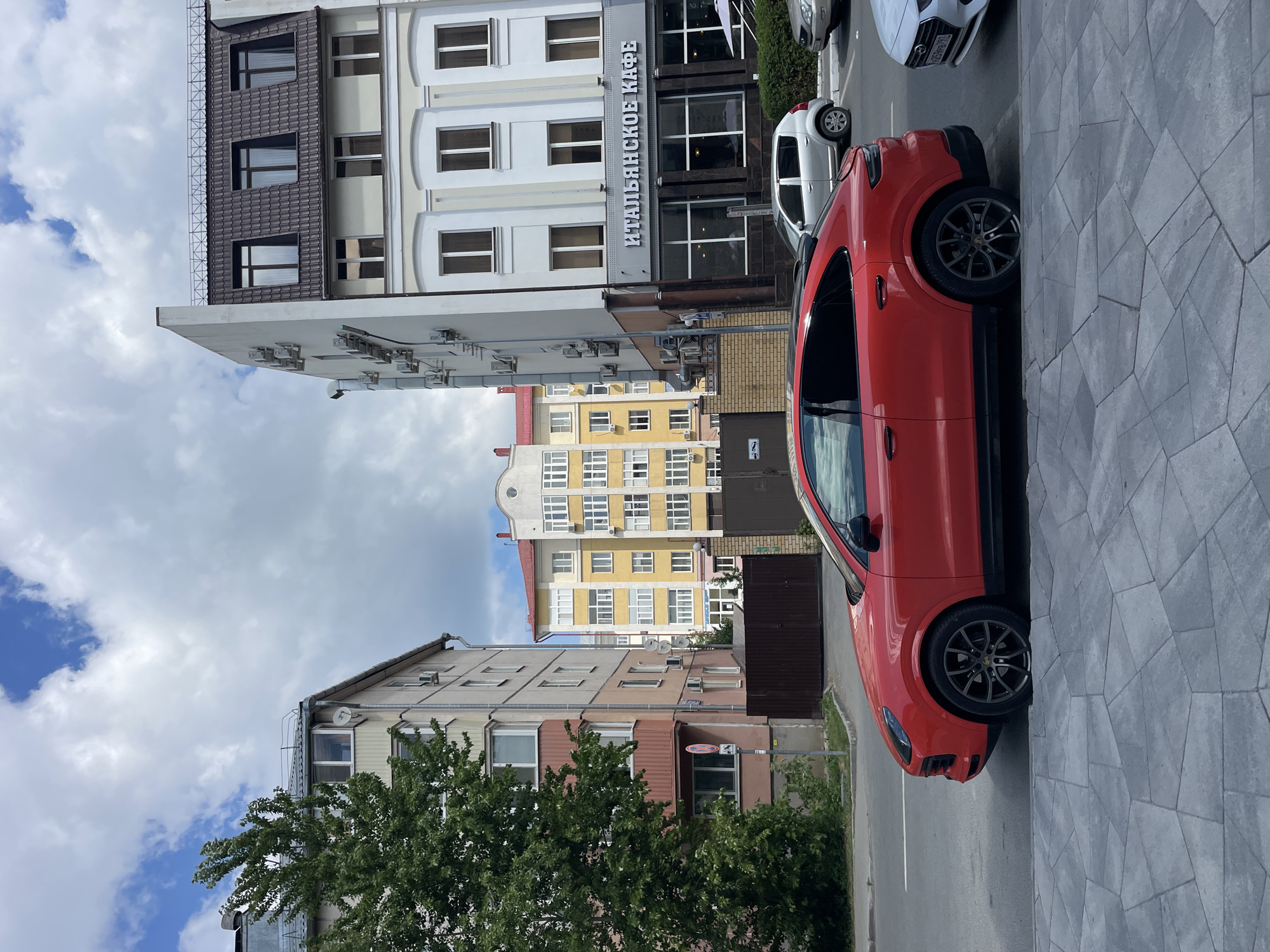 This is a street-level, eye-level, medium shot of a red Porsche Cayenne parked on a city street in Kazan, Russia. The car is facing right and is angled slightly towards the camera. The car is parked on the right side of the street, parallel to the sidewalk. The sidewalk is made of gray, geometric paving stones.
In the background, there are several buildings. The closest building on the right is a light-colored, multi-story building with a dark tiled roof. The building has large windows and a sign that reads "ИТАЛЬЯНСКОЕ КАФЕ" (Italian Cafe) in large, white letters. Next to this building, further in the background, is a bright yellow building with white window frames. To the left of the yellow building is a light-colored, multi-story apartment building with balconies. A large green tree is in front of this building.
Other vehicles are visible on the street, including a silver car parked behind the red Porsche and a white car parked to the right of the red Porsche. The sky is partly cloudy with patches of blue sky. The overall impression is a typical urban street scene.