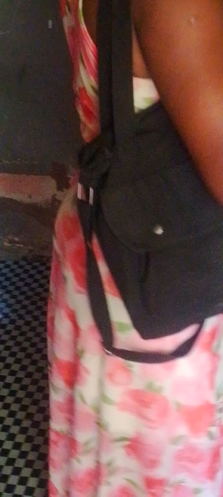The image shows a close-up view of a person wearing a floral dress and a dark-colored sling bag. The dress features a pattern of pink and white flowers with green accents. The sling bag is worn across the body. The background appears to be an indoor environment with a checkered floor in the lower left quadrant. The upper left and right portions of the background are darker and less defined, possibly indicating walls or furniture. There is no visible indication of the specific location within Jalingo, Nigeria.