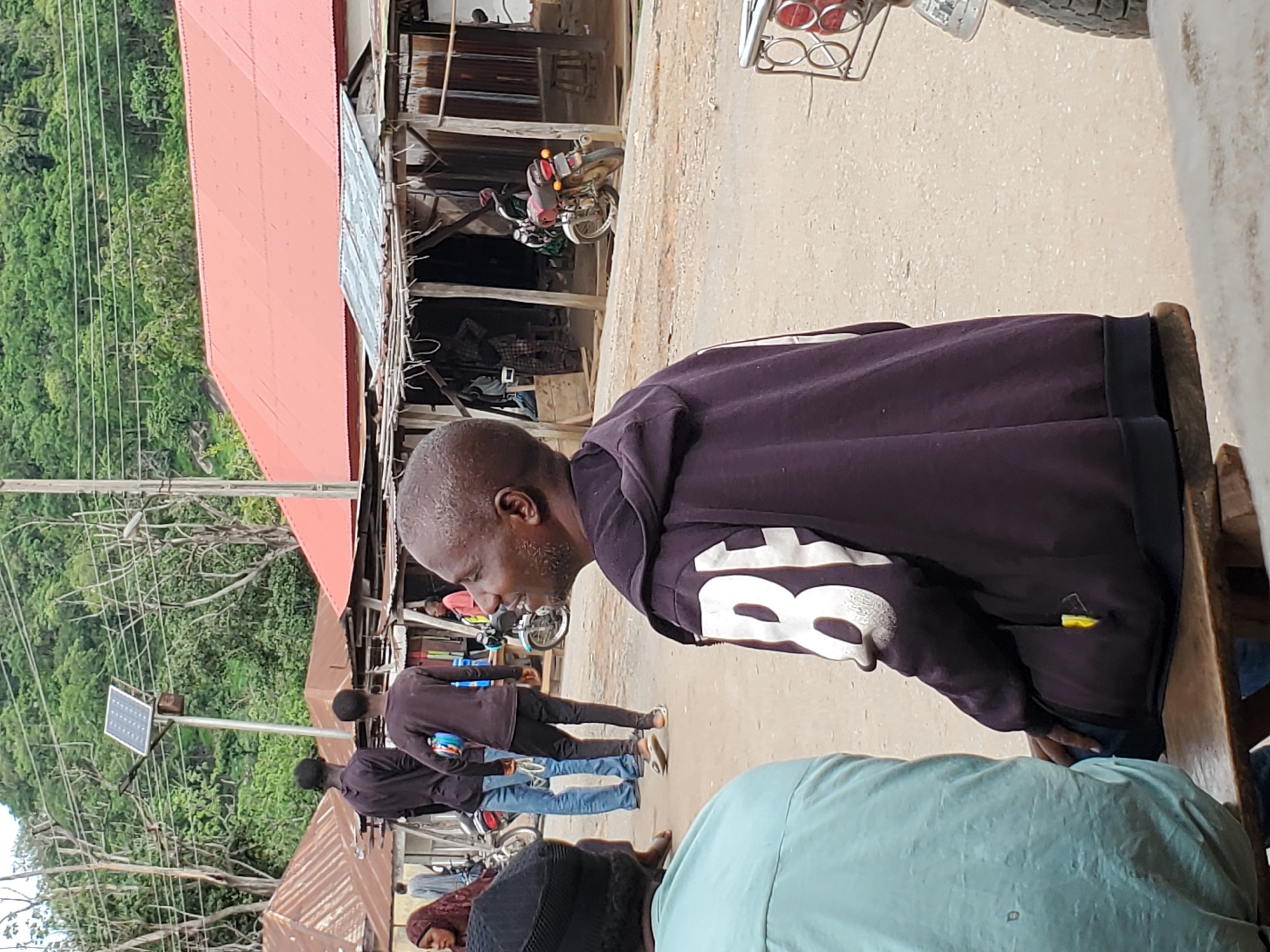 In Mararaba, Nigeria, the image captures a group of people outdoors near structures with red roofs. One man, wearing a dark purple hooded sweatshirt with the number "8" visible, is seated on a wooden structure. Other individuals are standing and walking in the background. A motorcycle is parked under one of the structures. Power lines and lush green vegetation are visible in the background. A solar panel is mounted on a pole nearby.