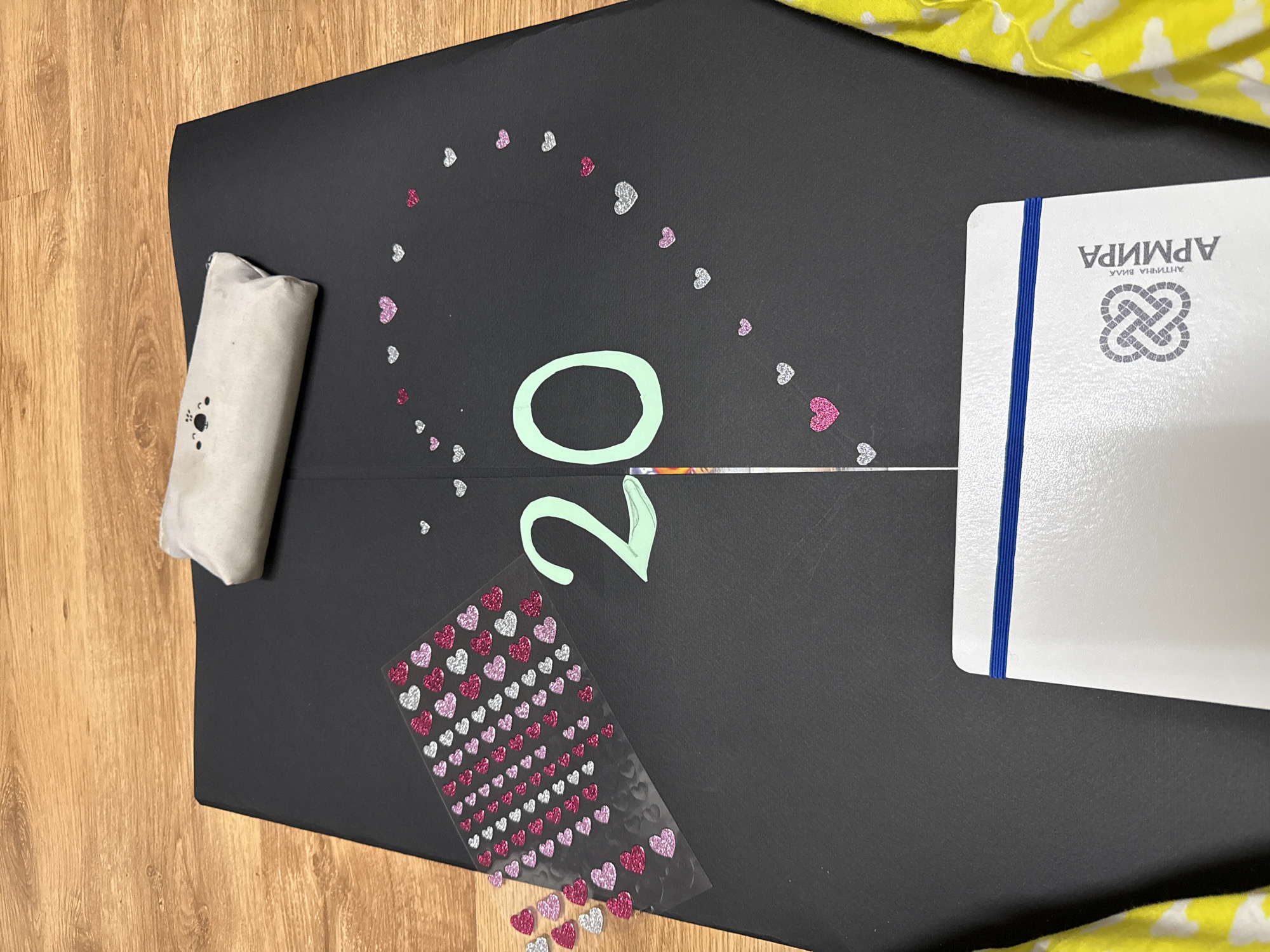 The image shows a dark surface decorated with hearts, a lightly colored rolled item, and a notebook. The numbers "20" are also present, seemingly cut out of paper. The notebook has the word "АРМИРА" written on it in Cyrillic. Given that "АРМИРА" is the name of a sports hall in Sofia, Bulgaria, it's plausible this image was taken in Sofia. The background includes a wooden surface and a yellow and white patterned fabric.