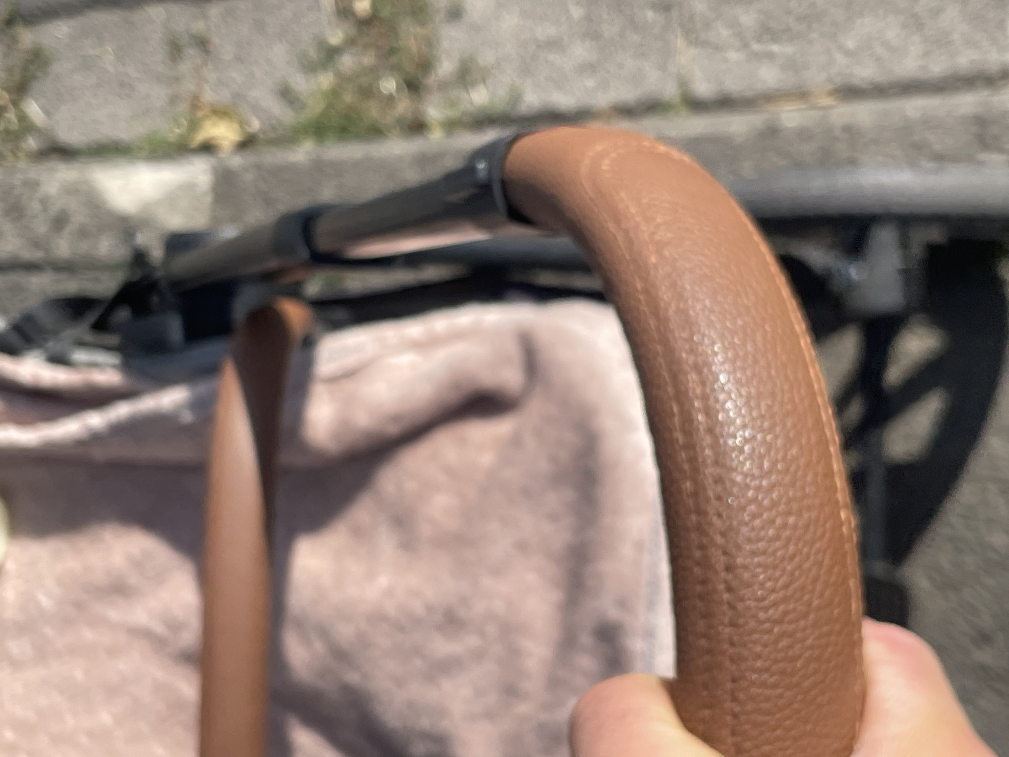 A close-up, slightly blurred, eye-level shot shows a person's hand gripping the brown, textured leather handle of a stroller. The stroller's fabric, a light pink or mauve color with a subtle pattern, is visible behind the handle. The stroller's black frame and wheel are also partially in view. The background consists of a gray paved surface, possibly a sidewalk, with some green foliage and fallen yellow leaves suggesting an outdoor setting in Neukirchen, Germany. The lighting indicates it is daytime, with sunlight creating some reflections and shadows.