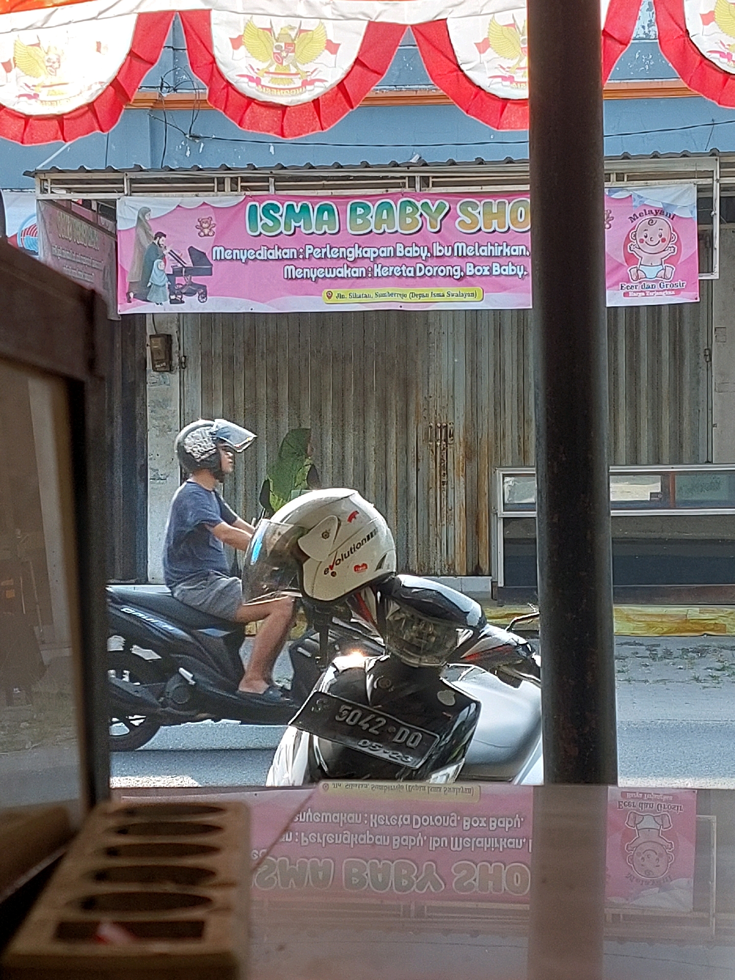A person is riding a motorcycle in front of a building in Sumuragung, Indonesia. The person is wearing a blue t-shirt and gray shorts. The motorcycle is black. There is a banner behind the motorcycle that says "ISMA BABY SHO". The banner also has a picture of a baby carriage and a teddy bear. The environment is a street with buildings and a wooden fence. The lighting is bright and sunny.