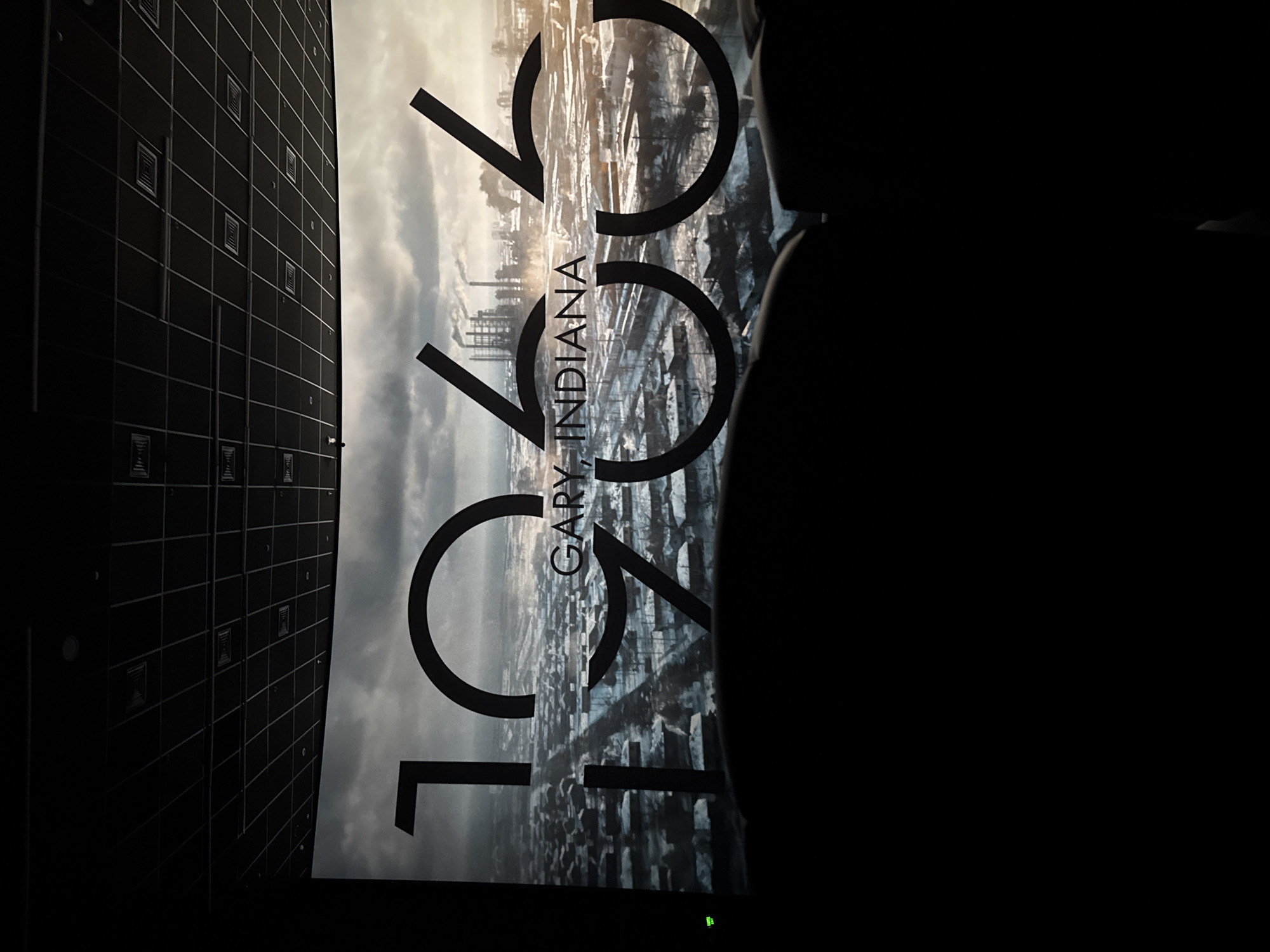 The image shows a screen displaying the text "1966, GARY, INDIANA." The background of the text features a cityscape with industrial elements like smokestacks and buildings. The photo appears to have been taken in a movie theater, with the grid-like pattern of a wall visible on the left and a silhouette of a person's head on the right.