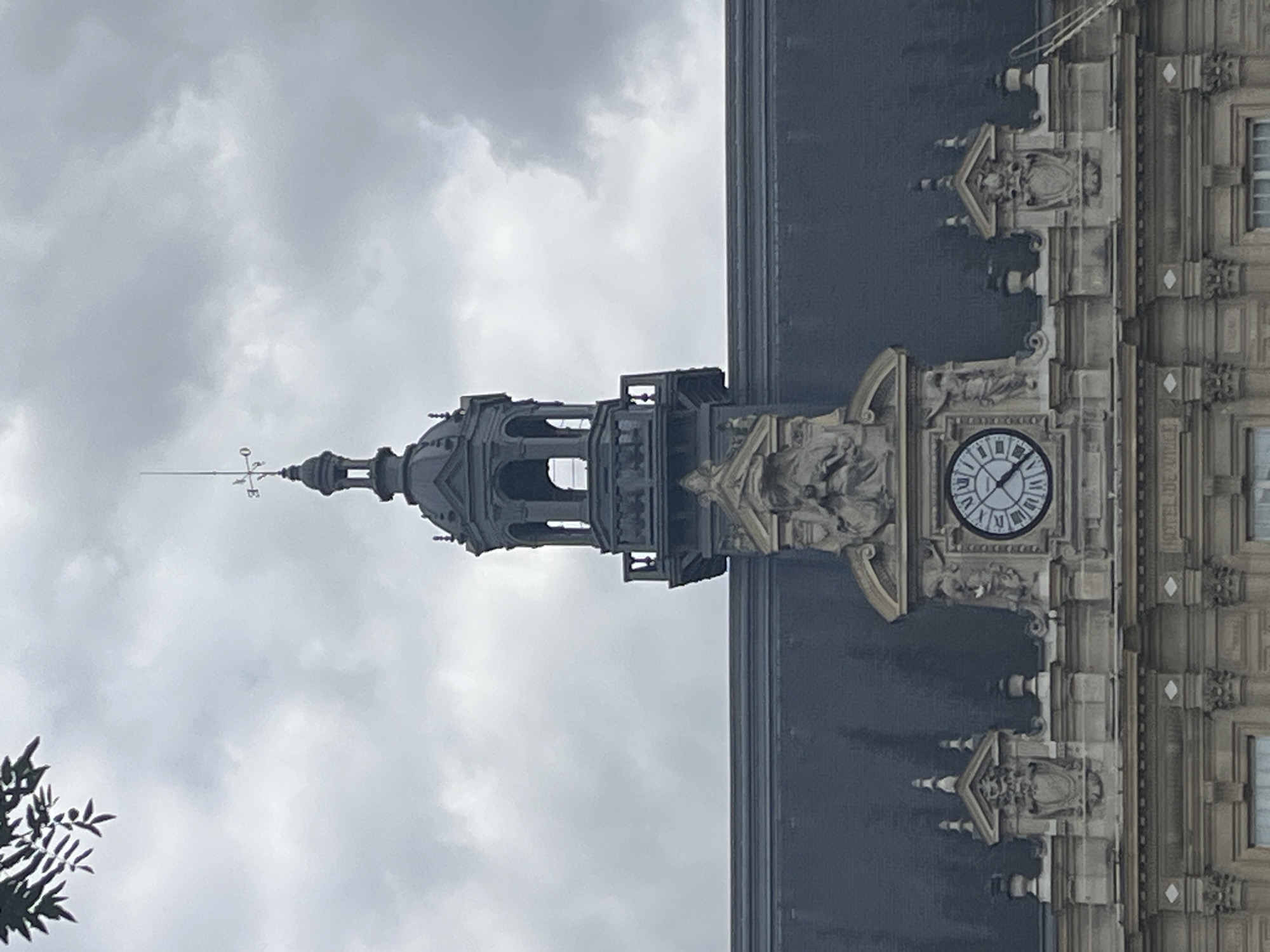 The image shows an exterior view of a building with ornate architectural details, including a clock tower. The building appears to be made of stone and features decorative carvings and moldings. A round clock face with Roman numerals is prominently visible on the clock tower. Above the clock, there is a smaller structure with open arches, and a weather vane extends from the top of the spire. The background is a cloudy sky, suggesting overcast weather. The scene is framed by some foliage on the left side of the image. The orientation of the image is vertical, with the clock tower pointing upwards. The setting appears to be a historic or grand building, possibly a public institution or a landmark. The overall mood is somewhat somber due to the cloudy sky. There is no discernible text in the image.