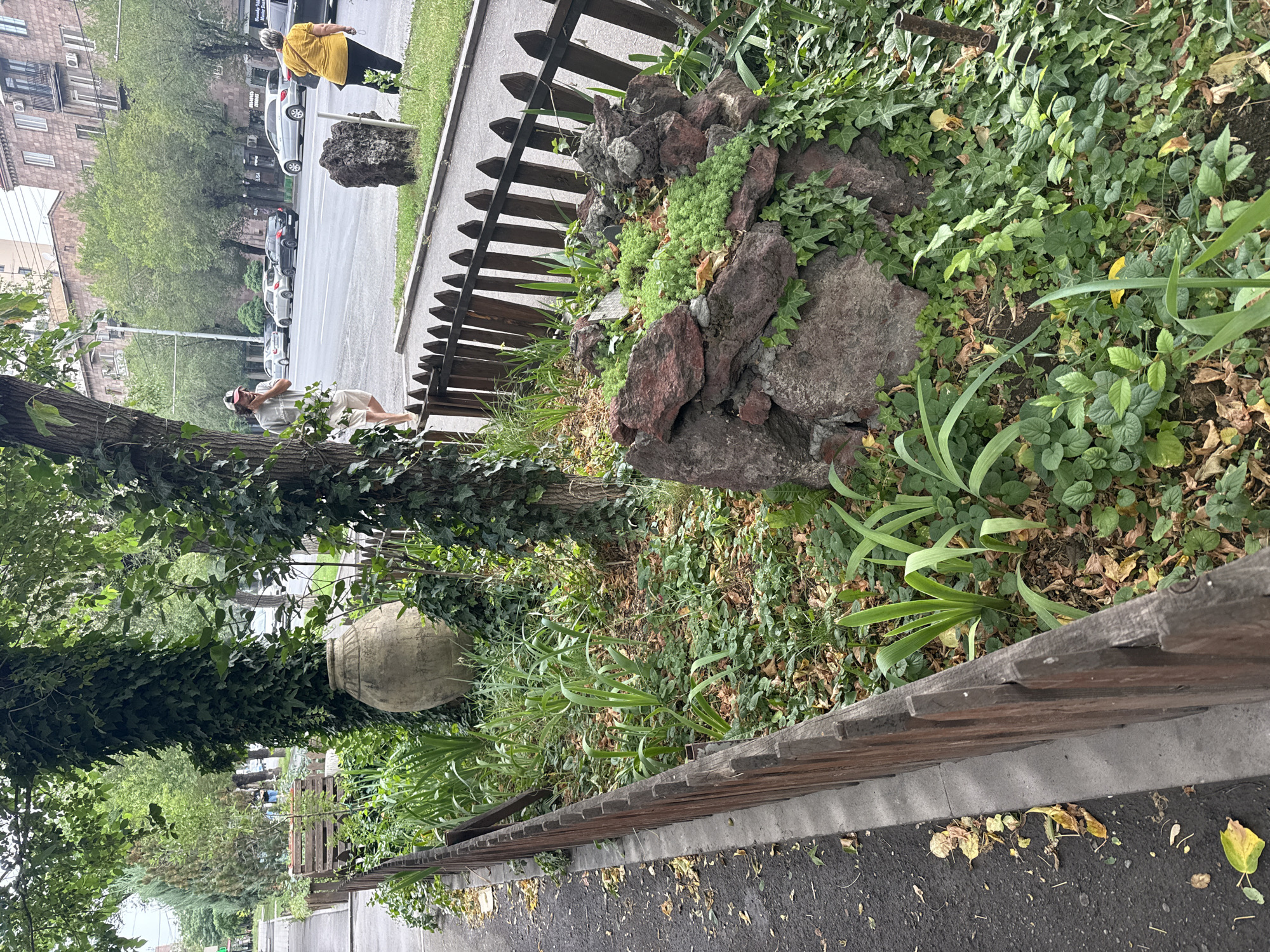 The image depicts a street scene in Yerevan, Armenia. In the foreground, a low wooden fence encloses a small garden area with lush greenery, fallen leaves, and a large, weathered clay amphora. A rocky mound with small green plants is also visible within the garden. Beyond the fence, a paved sidewalk runs alongside a street with several cars parked and driving. On the sidewalk, a man wearing a baseball cap and shorts is walking, and a woman in a yellow shirt is standing. Buildings with balconies line the street in the background. The overall scene suggests a moderately urban environment with patches of greenery.