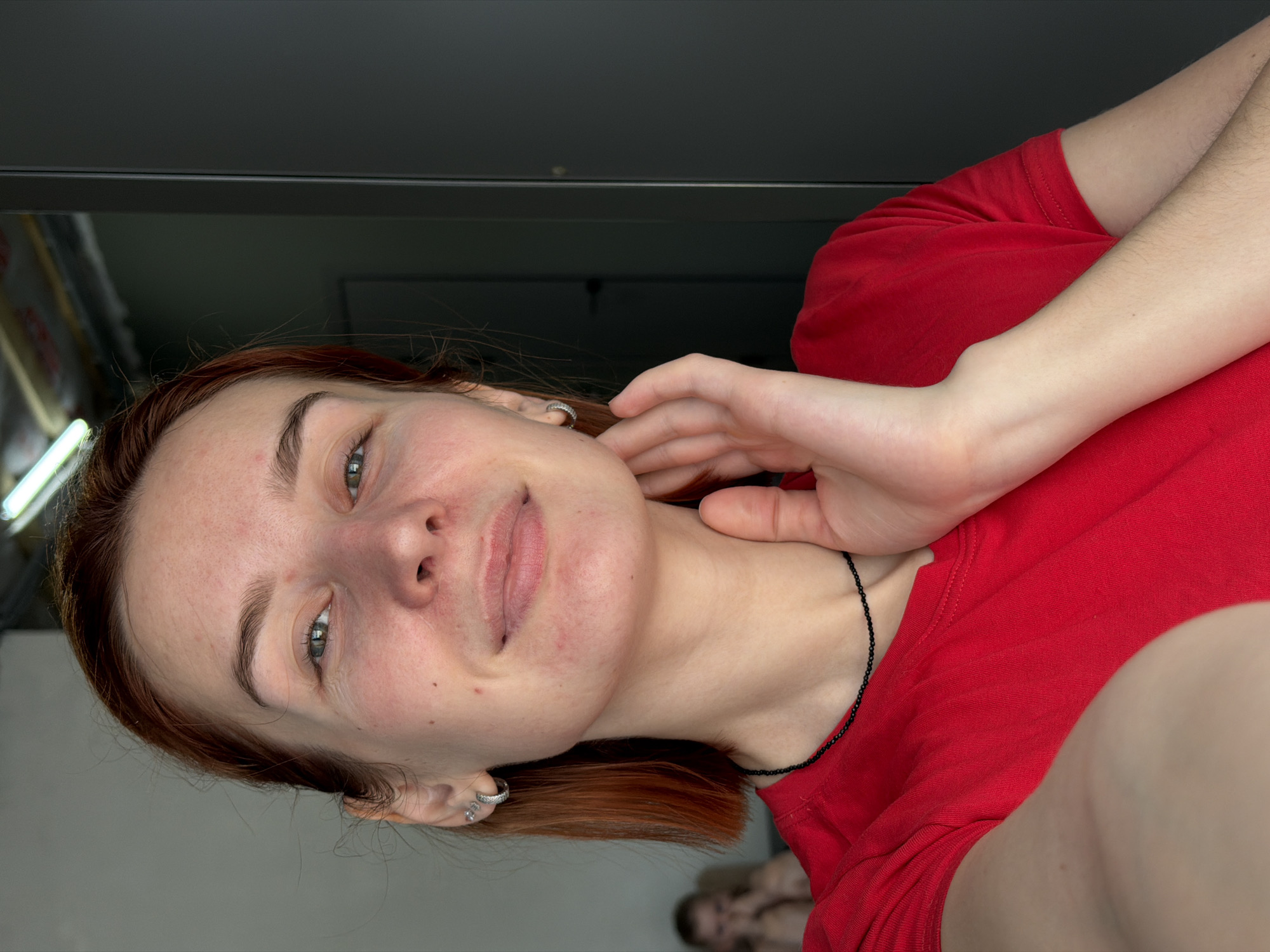 The image presents a close-up, front-facing portrait of an adult female taken indoors. The subject, positioned centrally, has reddish-brown hair, green eyes, and fair skin exhibiting minor redness and blemishes on her face. She wears a red short-sleeved t-shirt and a black beaded necklace. Her left ear is adorned with a silver hoop earring and two smaller stud earrings above it. Her right hand is raised, with fingers gently resting on the right side of her neck and jawline. Her expression is a slight smile, and she is looking directly at the camera.
In the blurred background to the left, a second individual, appearing to be a child or smaller adult with light-colored hair, is visible in a seated or crouching position, with their hands covering their face. Above the primary subject's head, a white fluorescent light fixture is partially visible against a ceiling that shows exposed structural elements. To the right of the primary subject, a dark gray panel, consistent with a door or cabinet, extends vertically, featuring a subtle seam and faint reflections. A dark door handle is dimly visible further in the background on the right side of the frame. The specific location within the city of Chaikovsky, Russia, is not identifiable from the visual information provided in the image.